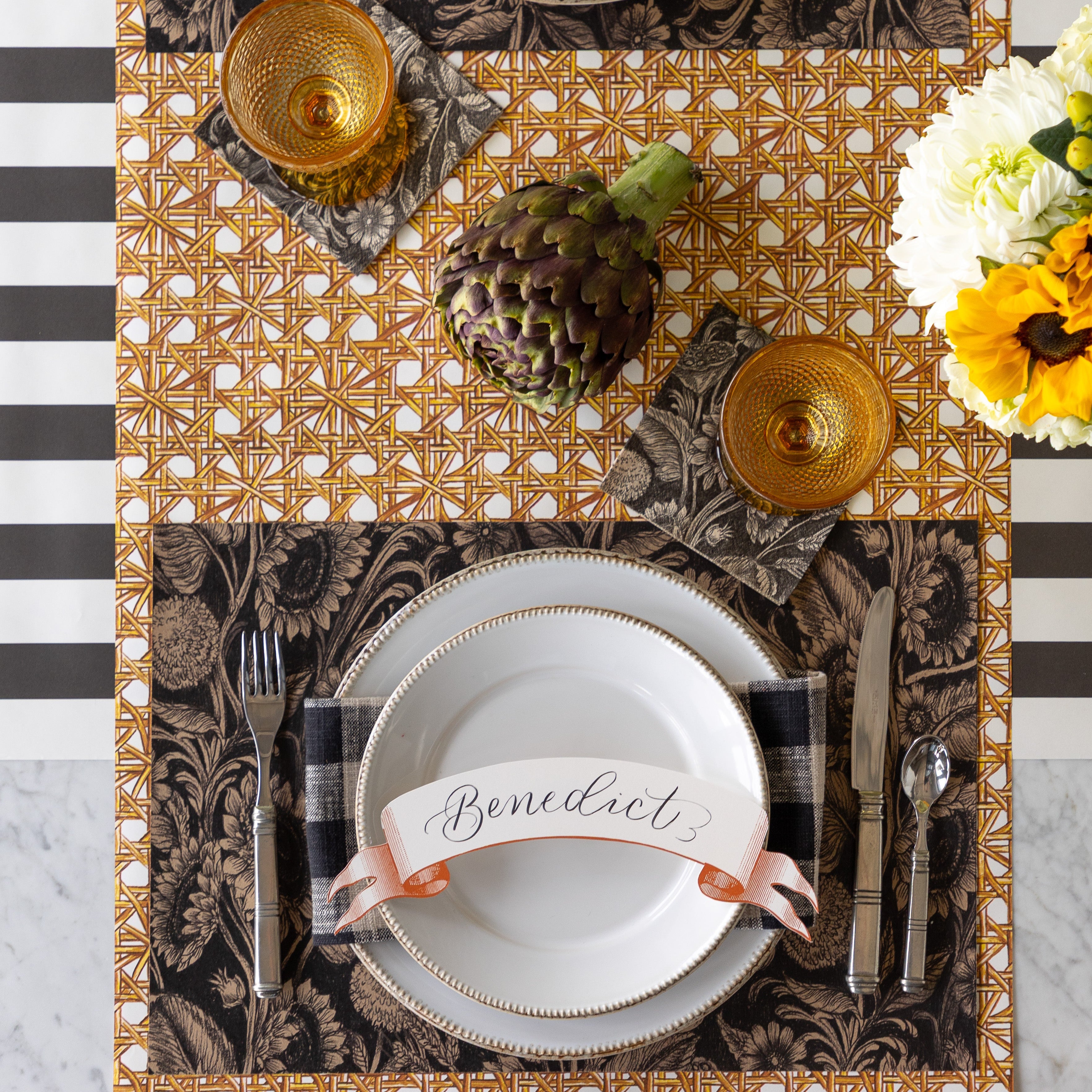A table setting with an artichoke centerpiece, featuring the Sunflower Field Placemat under white dinner plates with the Orange Classic Banner Table Accent reading "Benedict" atop, Vintage Sunflower Cocktail Napkins under orange glasses, Rattan Weave Runner rolled vertically and Black Classic Stripe Runner rolled horizontally underneath.