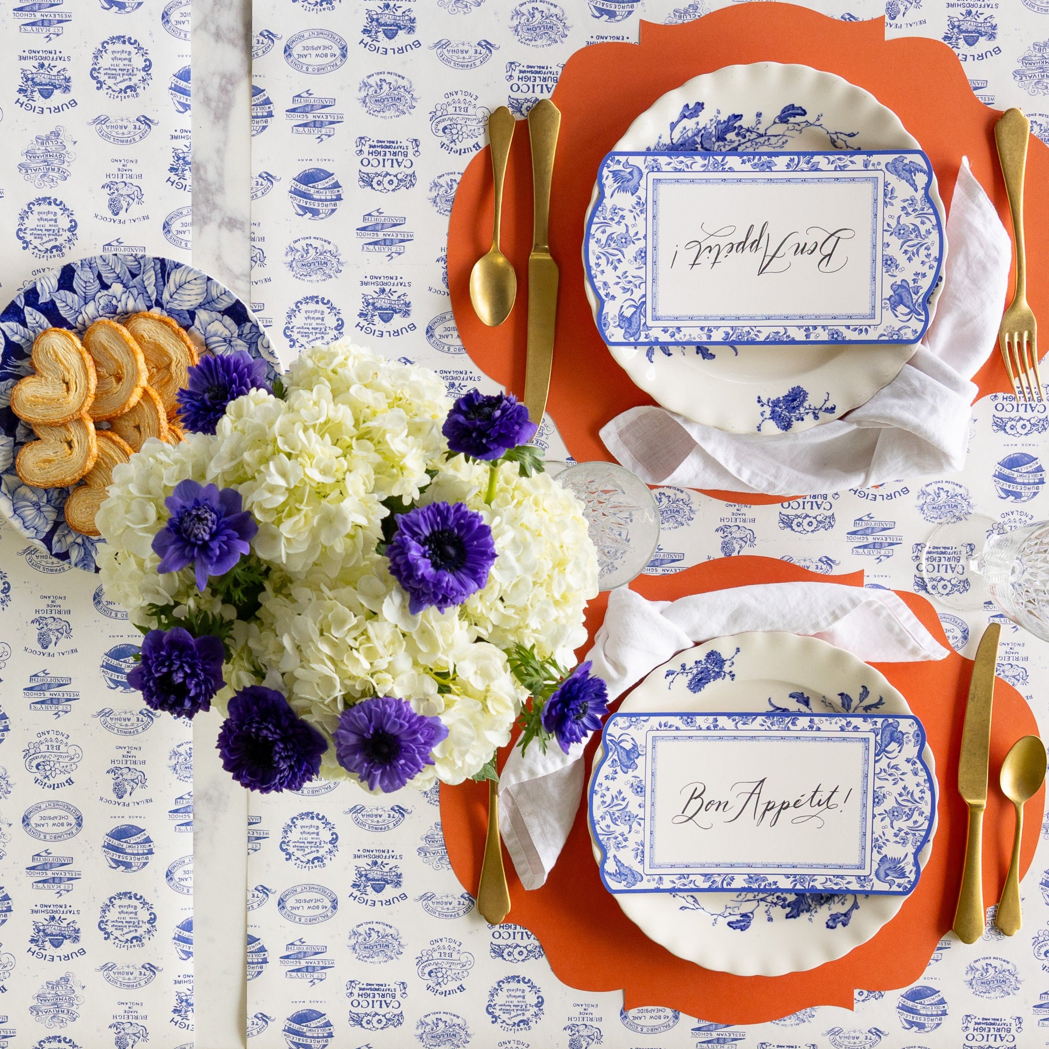 A table set for two with blue and white plates on Die-cut Orange French Frame Placemats gold cutlery, a bouquet of white and purple flowers, and plate of cookies on the Burleigh Heritage Runner. The plates feature Blue Regal Peacock Table Accents reading "Bon Appétit!" atop.