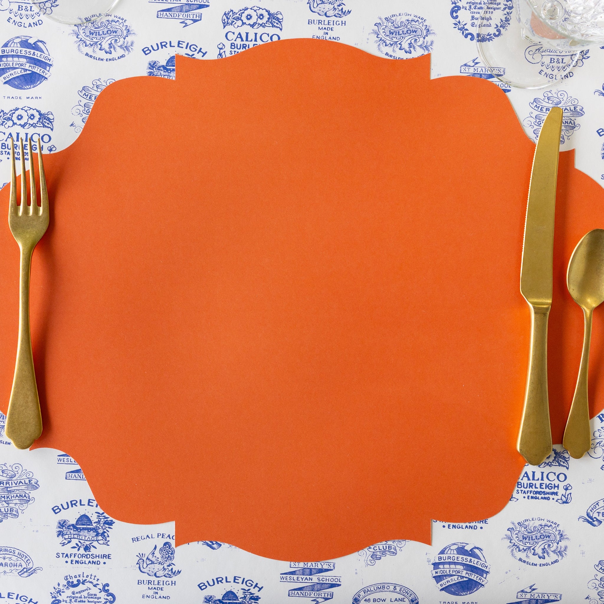 The Die-cut Orange French Frame Placemat with gold flatware atop, on the Burleigh Heritage Runner.