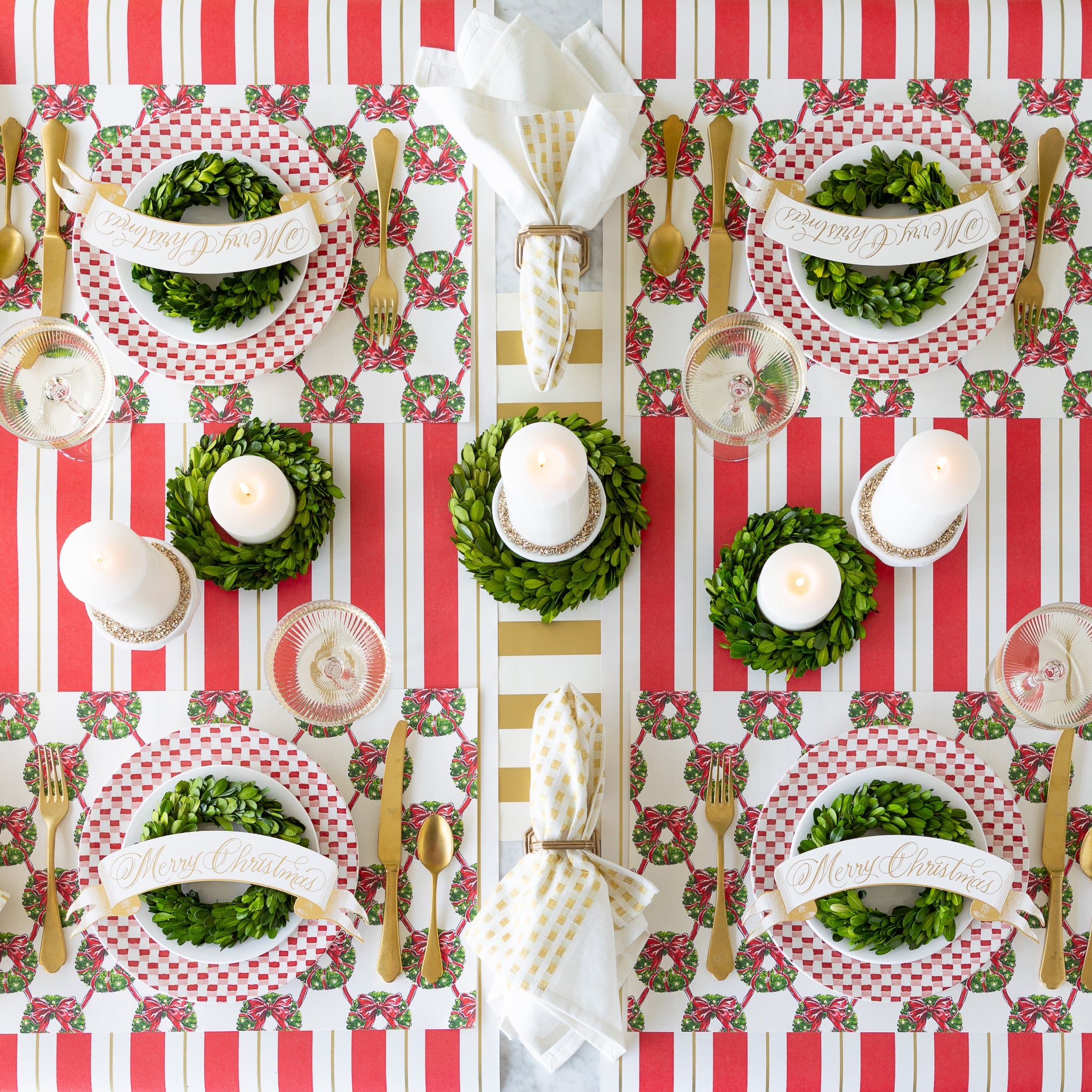 A festive table setting featuring Holiday Wreath Lattice Placemats, red-and-white plates adorned with wreaths and Merry Christmas Banner Table Accents, candles, and gold accents on the Antique Red Striped Runner.