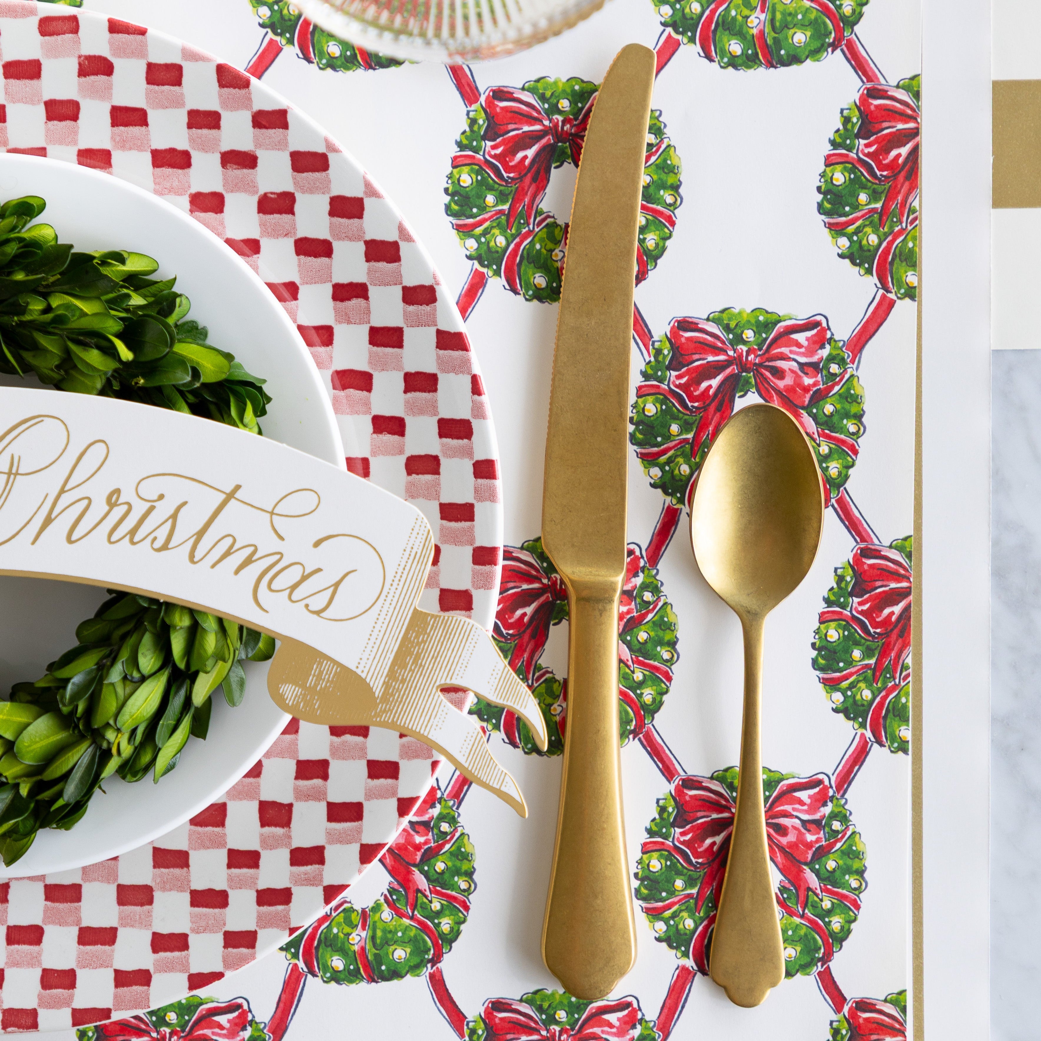 The Holiday Wreath Lattice Placemat under an elegant place setting with gold flatware, a red-and-white checkered plate, boxwood wreath, and Merry Christmas Banner Table Accent.