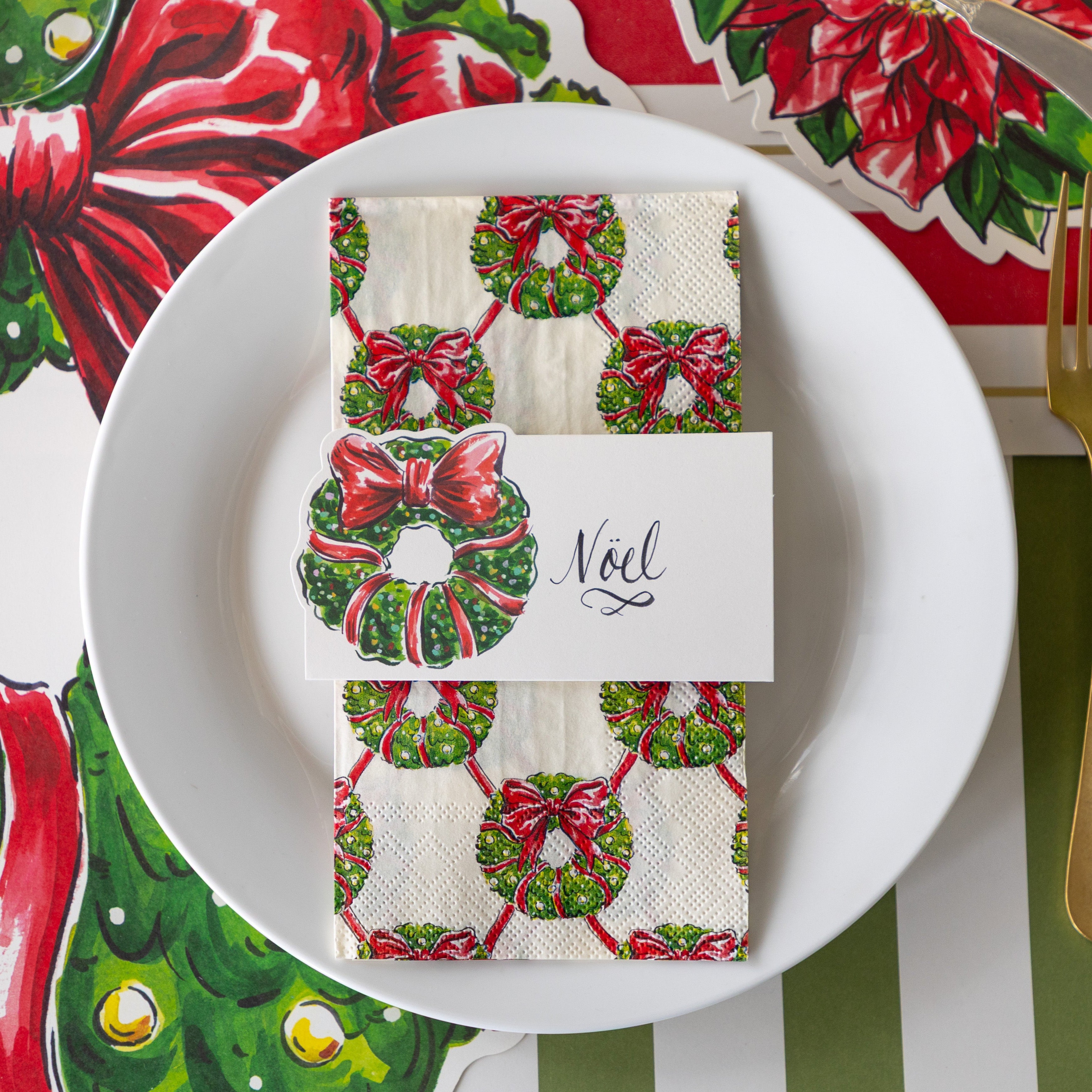 A place setting with the Holiday Wreath Place Card reading "Noel", atop the Holiday Wreath Guest Napkin and white plate, on the Die-cut Holiday Wreath Placemat.