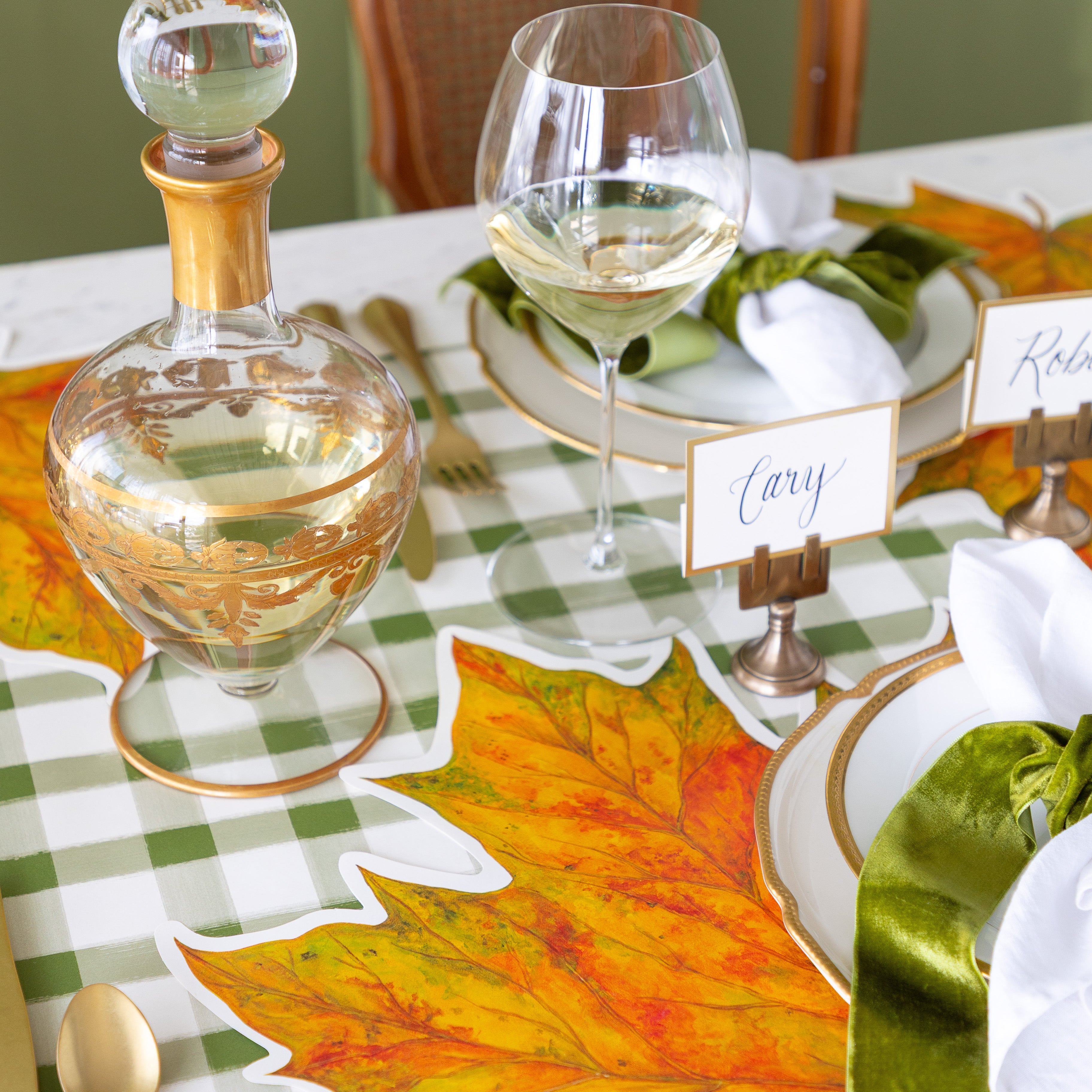 An elegant place setting featuring the Die-cut Fall Leaf Placemat, gold rimmed plates, the Gold Frame Place Card held by the Brass Place Card Holder, white napkins tied by a green velvet ribbon and the Green Painted Check Runner underneath.