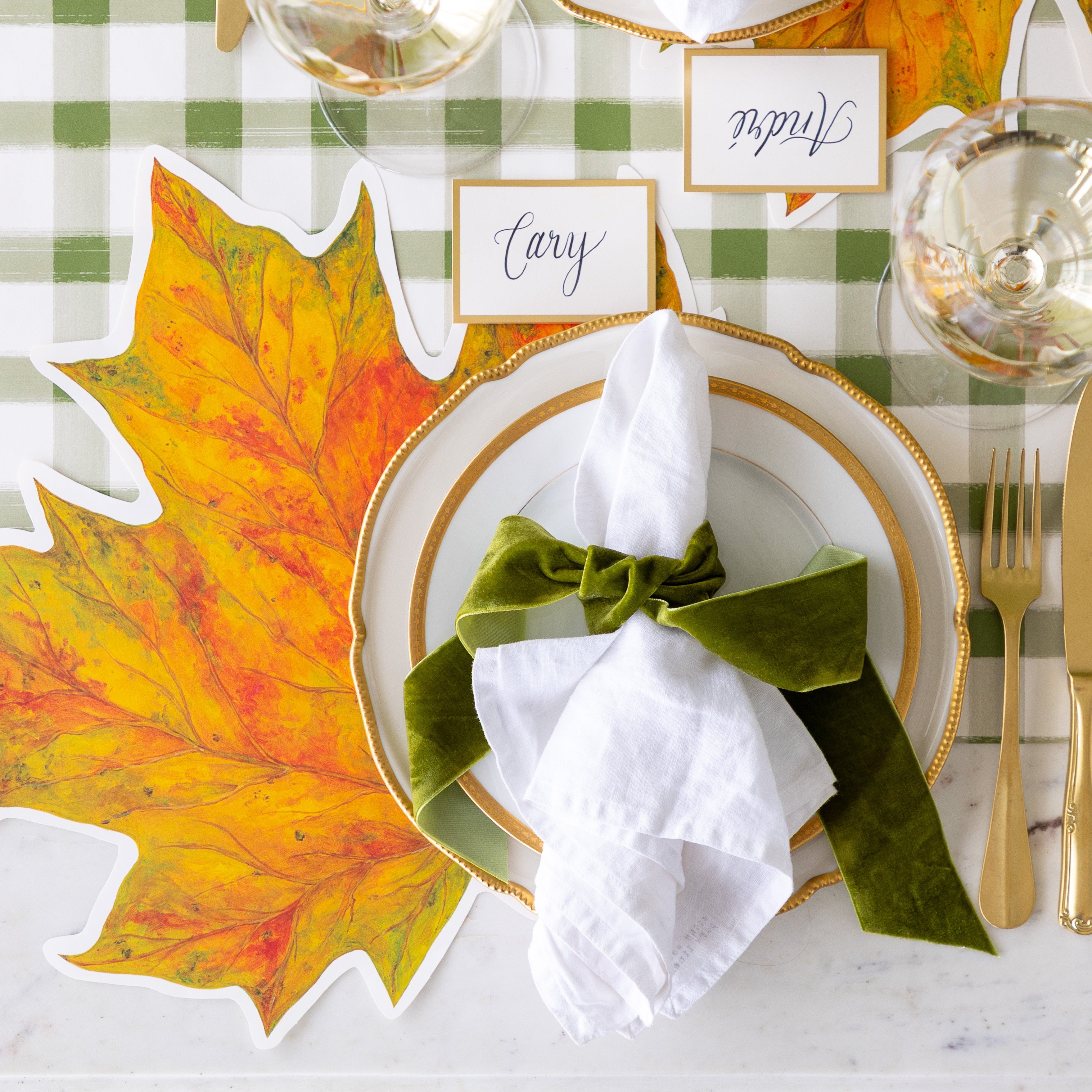 An elegant place setting featuring the Die-cut Fall Leaf Placemat, gold rimmed plates, the Gold Frame Place Card reading "Cary", white napkins tied by a green velvet ribbon and the Green Painted Check Runner underneath.