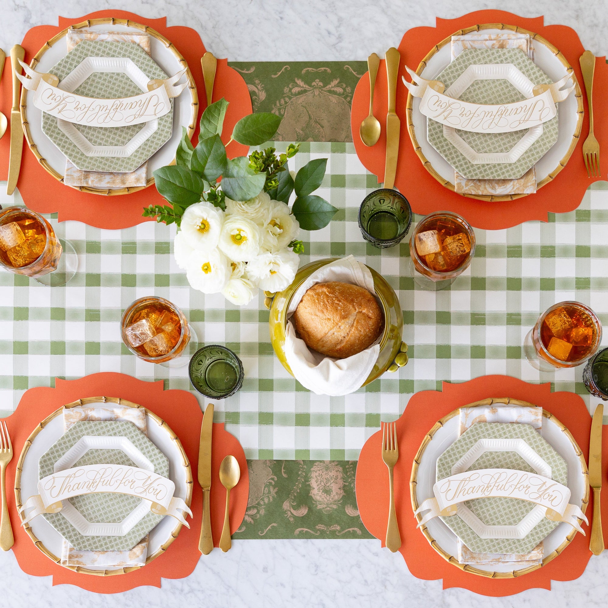 A fall table setting with a floral centerpiece and loaf of bread, featuring the Die-cut Orange French Frame Placemats, vintage dinnerware, floral napkins, gold flatware, and iced tea on the Moss Painted Check and Moss Fable Toile Runners.