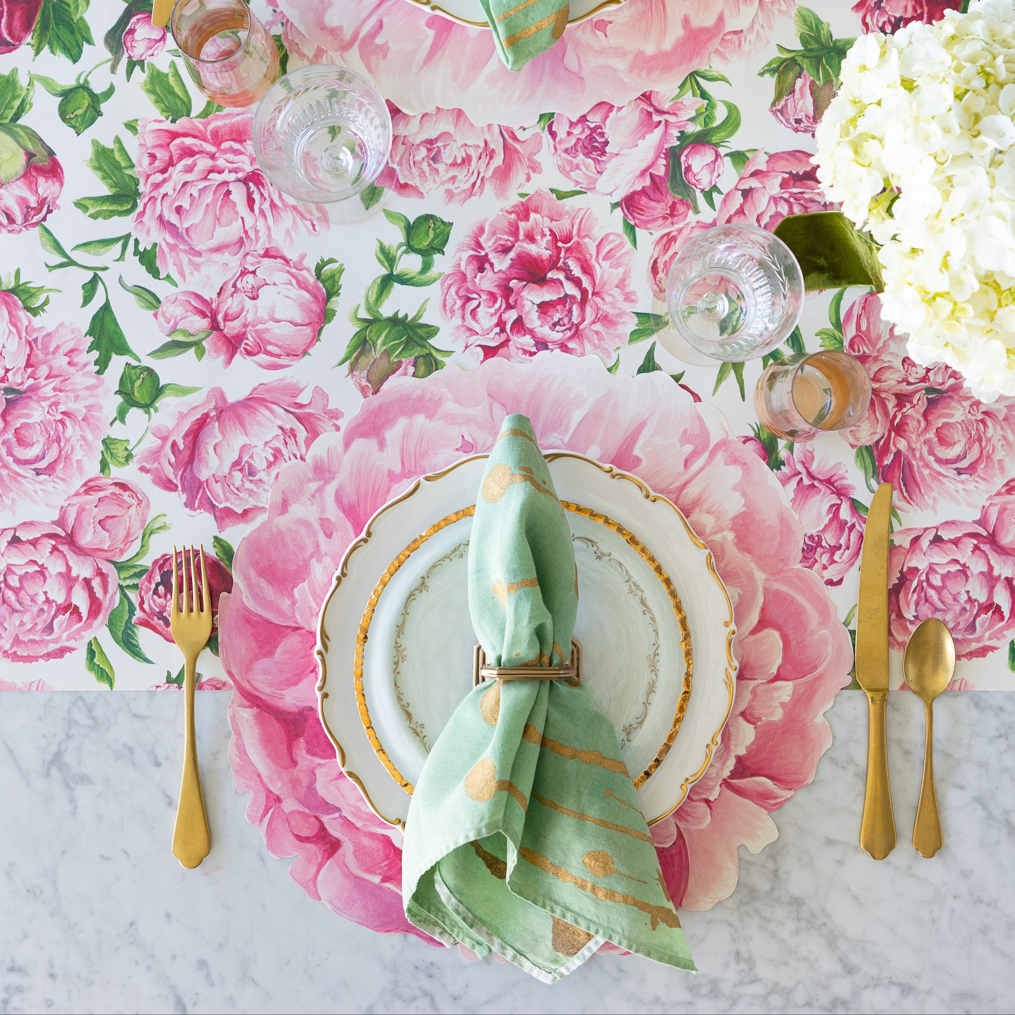 A table setting with the Peonies in Bloom Runner, Die-cut Peony Placemat, decorative plates, Peonies Place Card, marbled napkin and gold flatware.