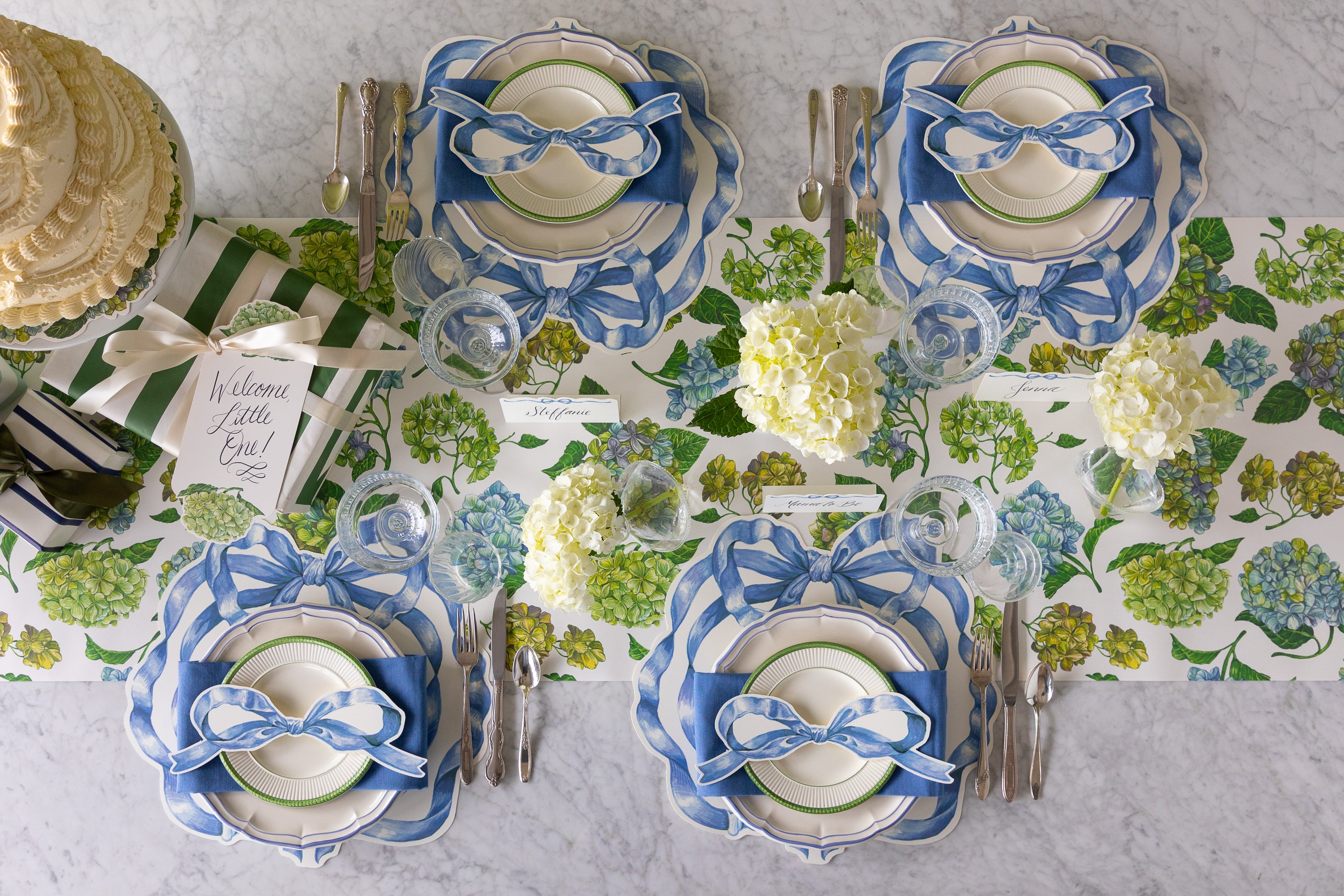 Set table with Hydrangeas, Die-cut Heirloom Blue Bow Placemats, table accents and place cards, vintage flatware, a cake and baby gift on the Hydrangea Runner.