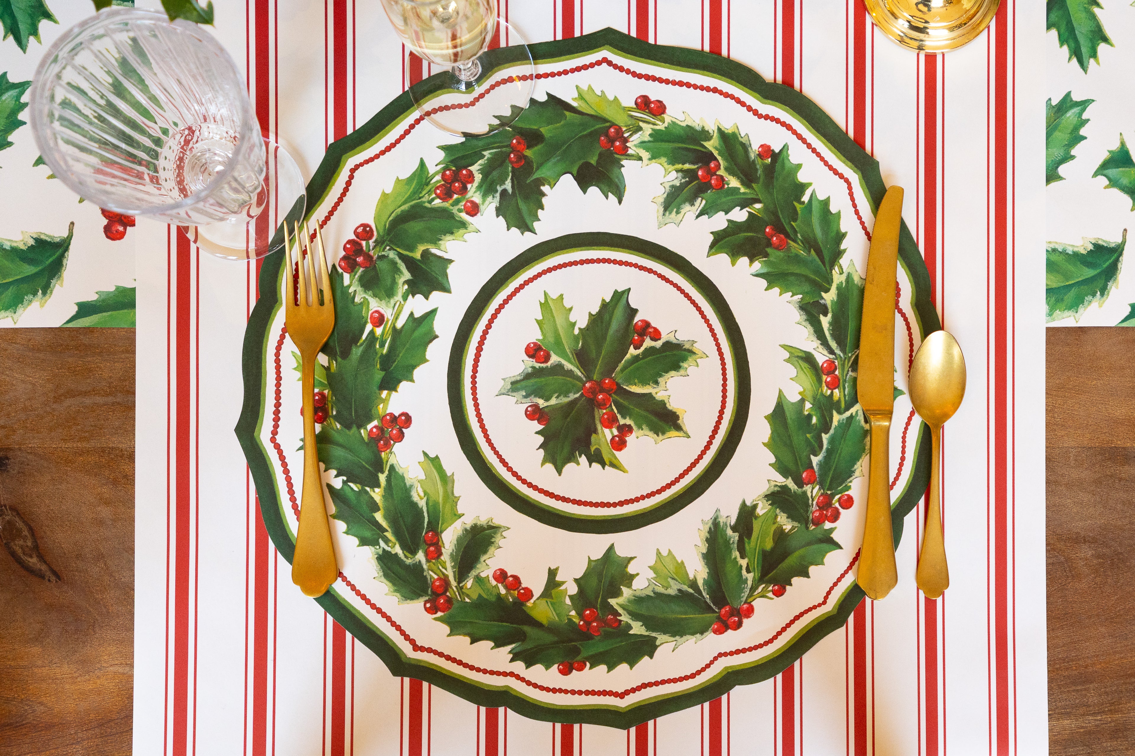Die-cut Holiday Holly China Placemat on a red striped table runner with gold cutlery on it.