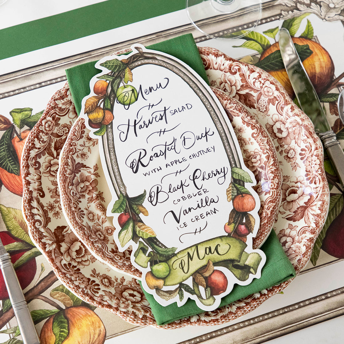A Heirloom Apples Table Card with a menu written inside the frame and &quot;Mac&quot; written on the ribbon, resting on the plate of an elegant place setting.