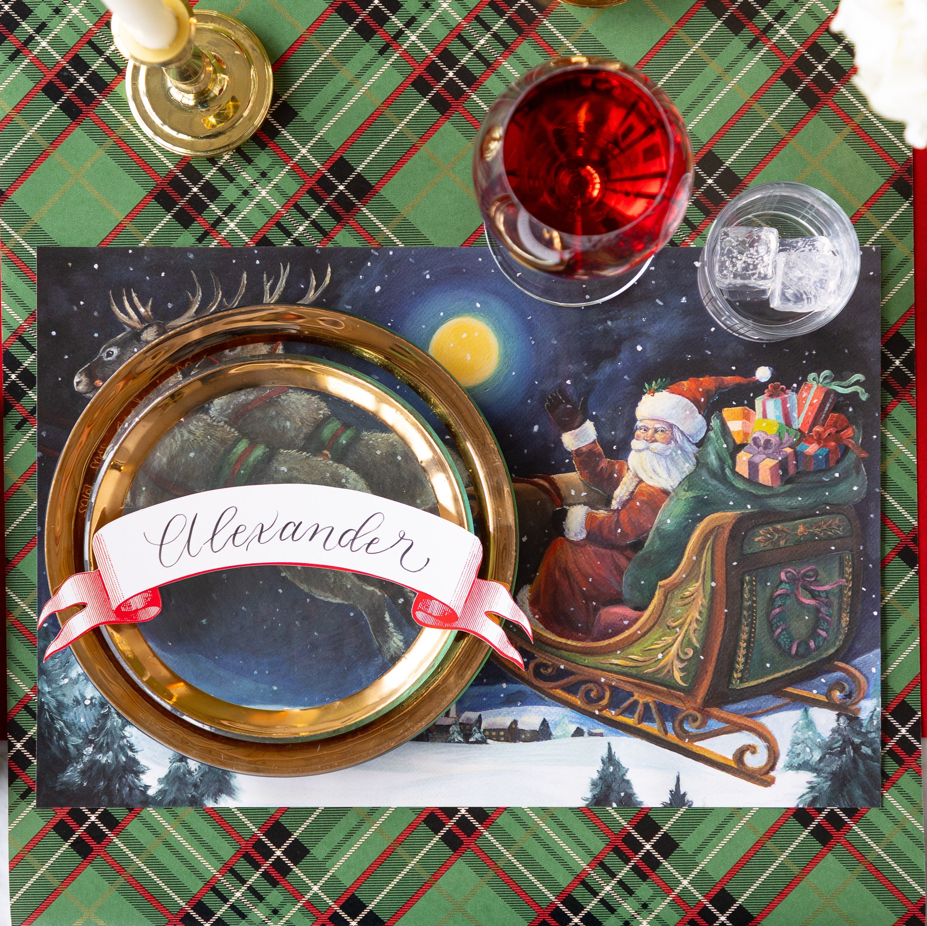 A Christmas place setting featuring the Sleigh Ride Placemat, gold rimmed dinner plates with the Classic Red Banner Table Accent reading "Alexander" atop, a glass of wine, a candle and Green Plaid Runner underneath.