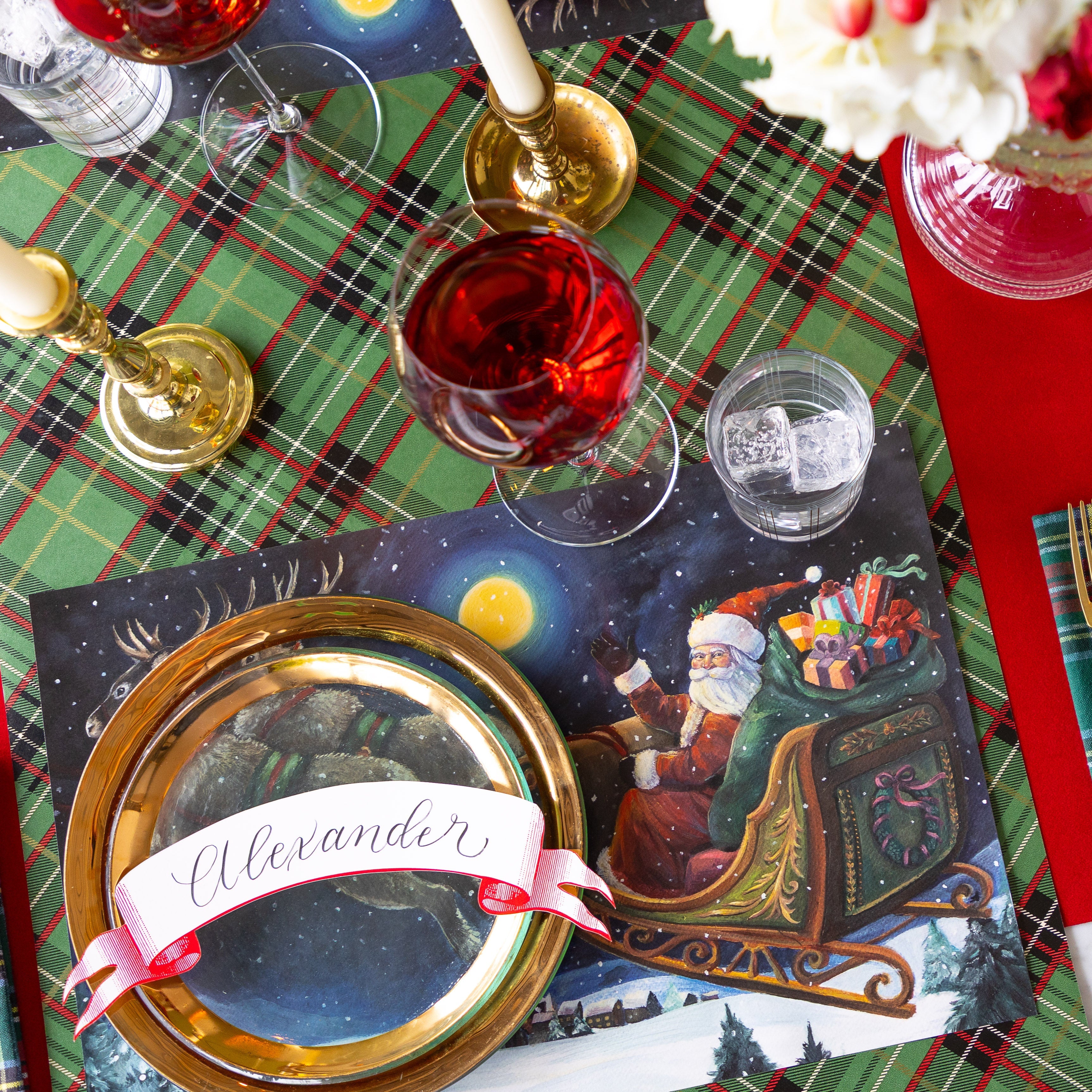 A Christmas place setting featuring the Sleigh Ride Placemat, gold rimmed dinner plates with the Classic Red Banner Table Accent reading "Alexander" atop, a glass of wine, candles and Green Plaid Runner underneath.