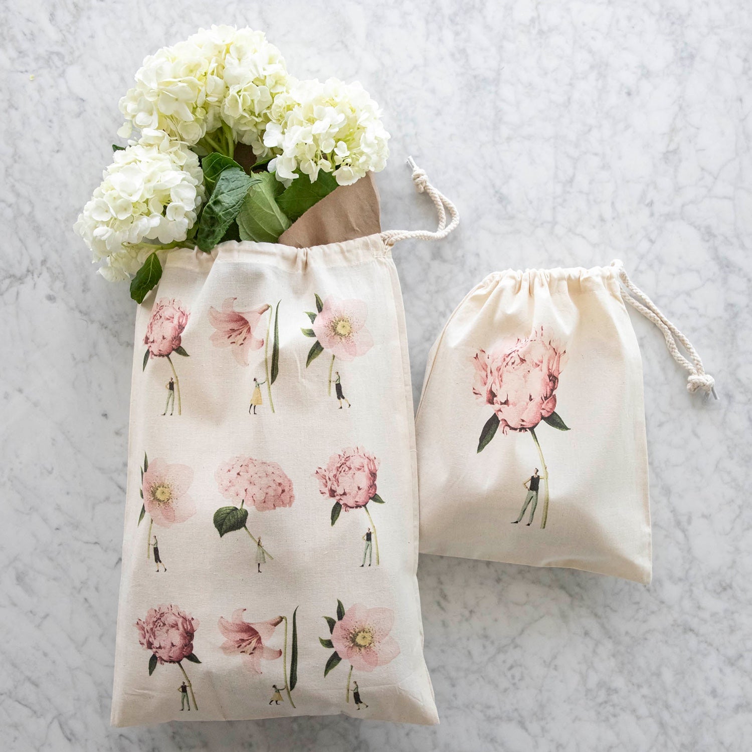 Two of the Laura Stoddart Drawstring Bags - the Medium Pink Flowers Bag and the Small Pink Peony Bag, both resting on a white table.