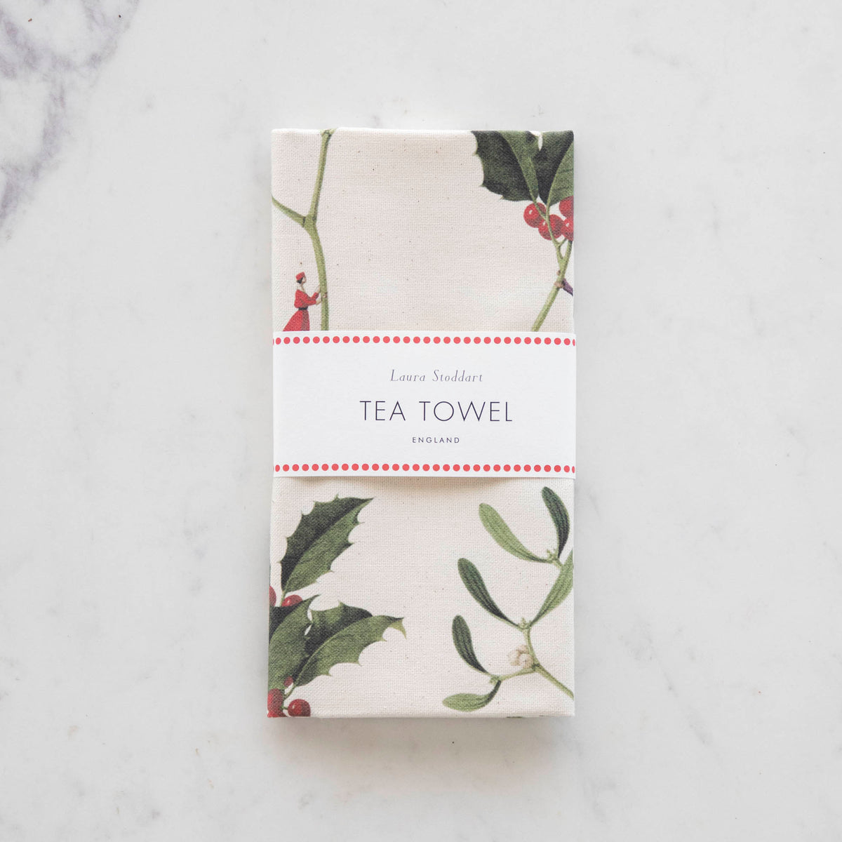 The Laura Stoddart Holly & Mistletoe Tea Towel folded with a white band, trimmed with red dots around it, and the words "Laura Stoddart", "Tea Towel" and "England" on it.
