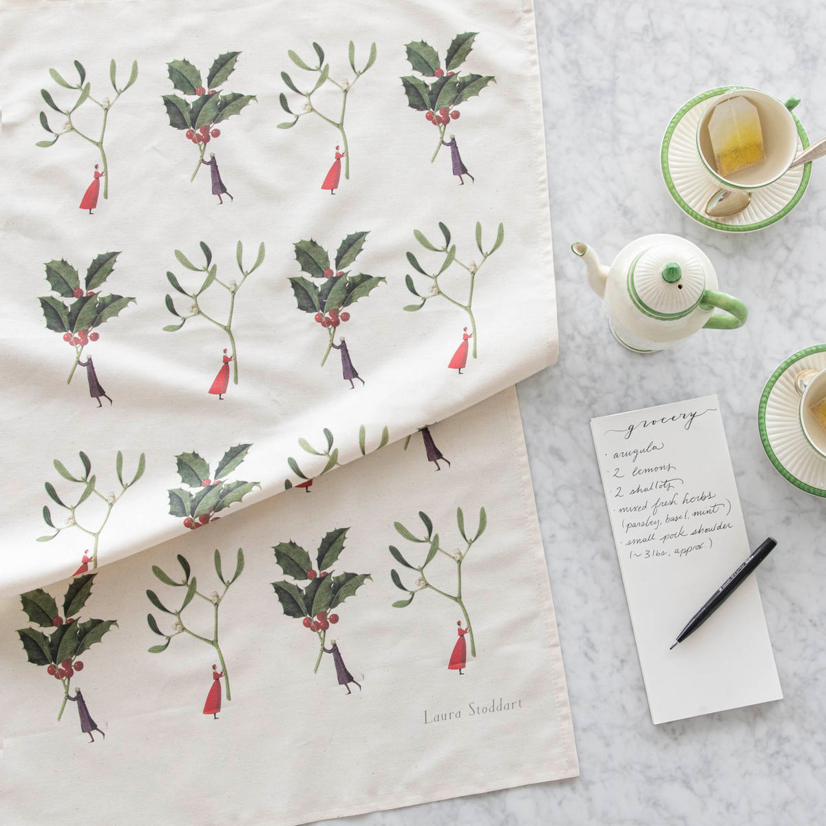 Laura Stoddart Holly & Mistletoe Tea Towel next to a notepad with a grocery list written on it, tea cups and a creamer pot.