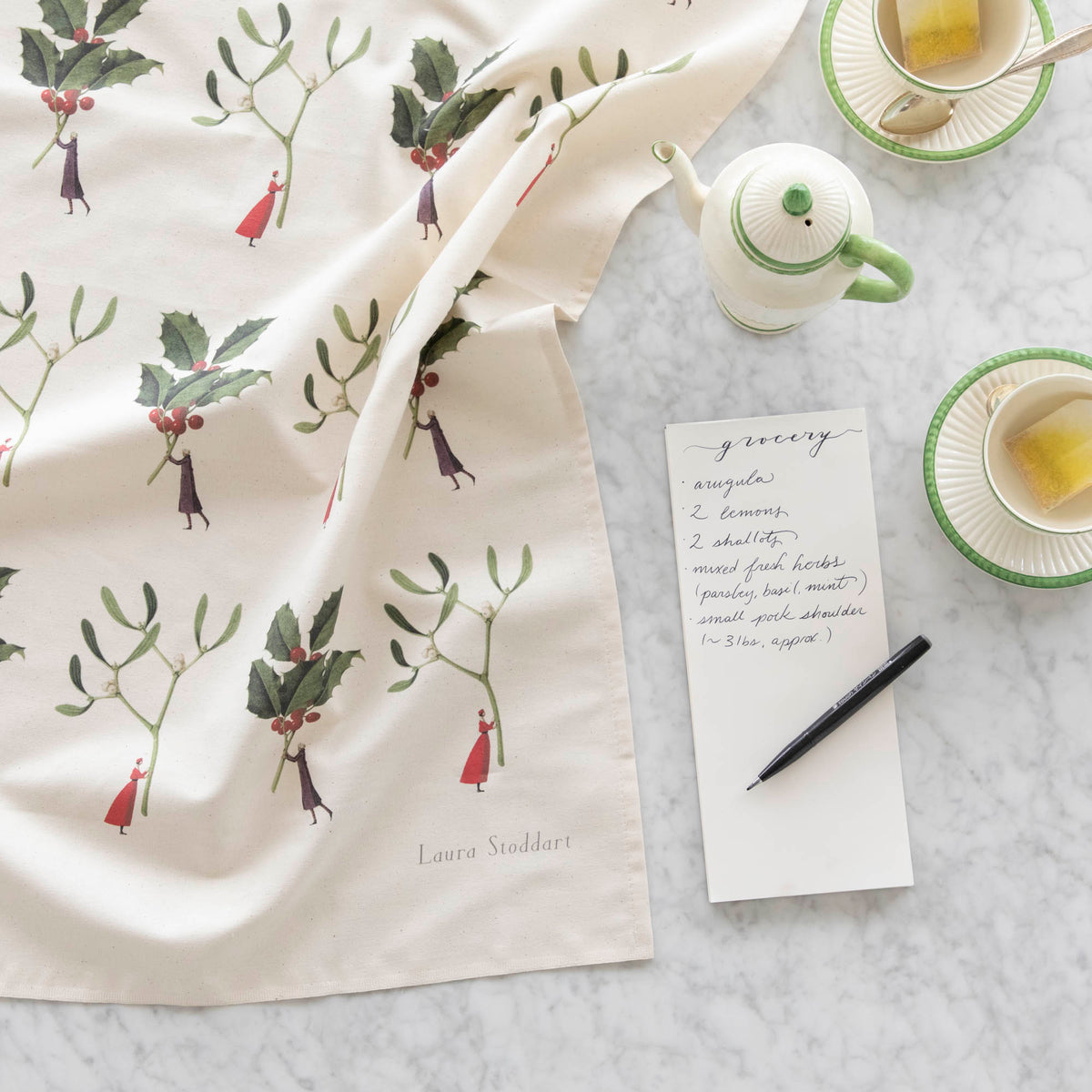 Laura Stoddart Holly & Mistletoe Tea Towel next to a notepad with a grocery list written on it, tea cups and a creamer pot.