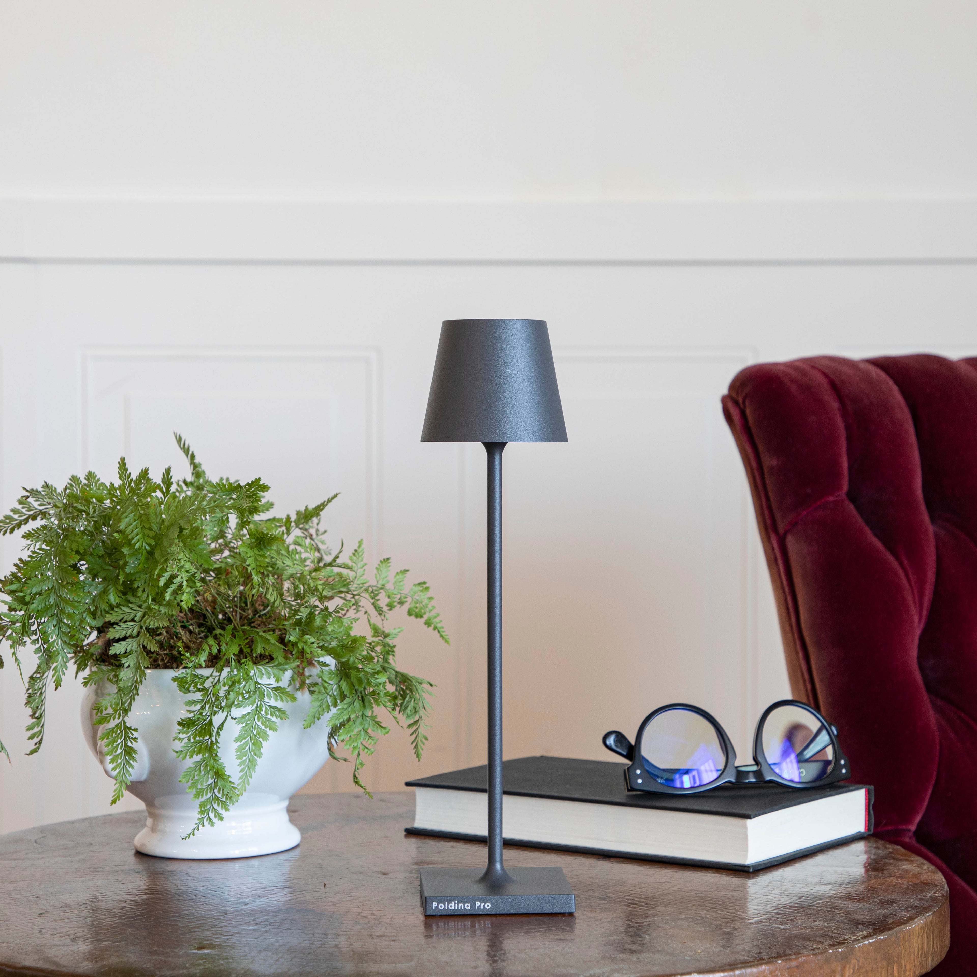 A Zafferano Dark Grey Micro Cordless Lamp on a table next to a book and a plant.
