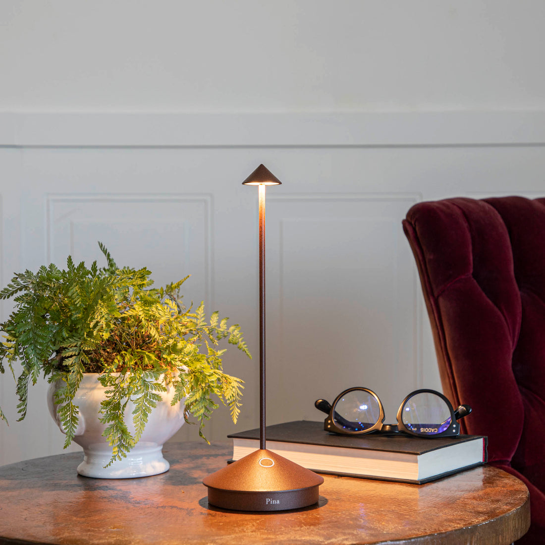 A modern Zafferano Rust LED cordless desk lamp with 3 color temperatures illuminates a cozy reading nook with a plant, glasses, and a book.