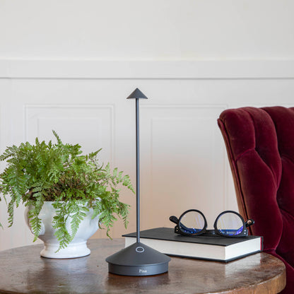 A Dark Grey LED Cordless Lamp by Zafferano on a table next to a book and a plant.
