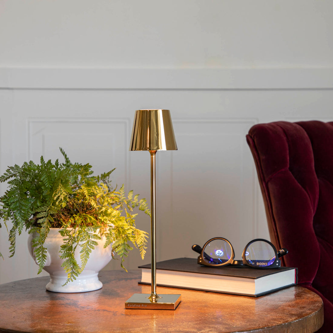 A Zafferano Glossy Gold Micro Cordless Lamp illuminates a corner featuring a potted fern, a closed book, and eyeglasses on a wooden side table.