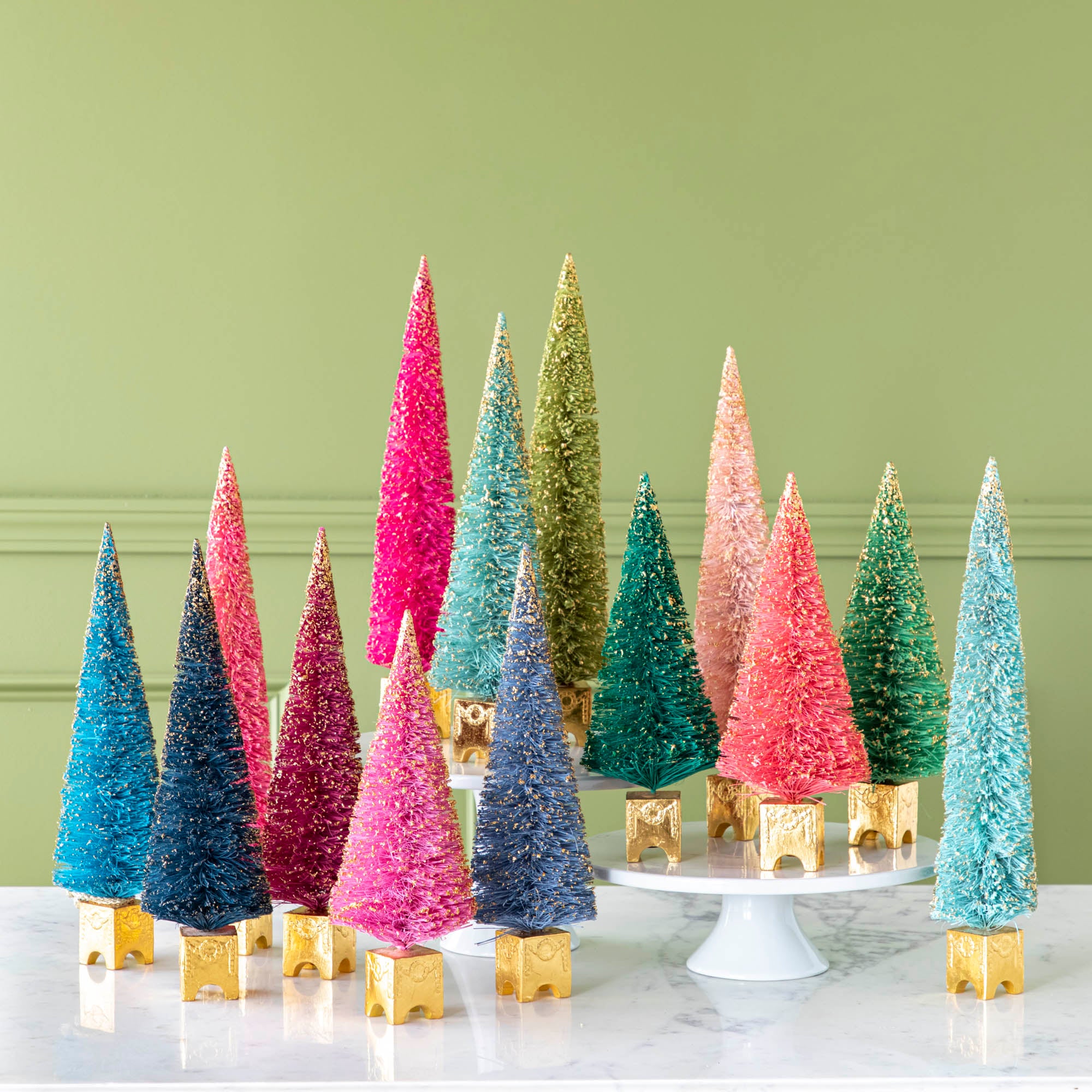 A collection of colorful artificial Christmas trees with gold leaf detailing, displayed on a white surface.