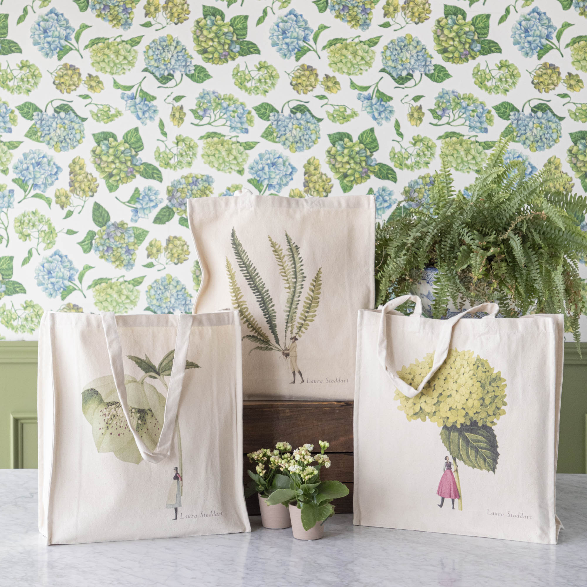 Three of the Heavyweight Canvas Tote Bags featuring green botanical artwork by Laura Stoddart arranged alongside potted plants against a hydrangea wallpaper background.