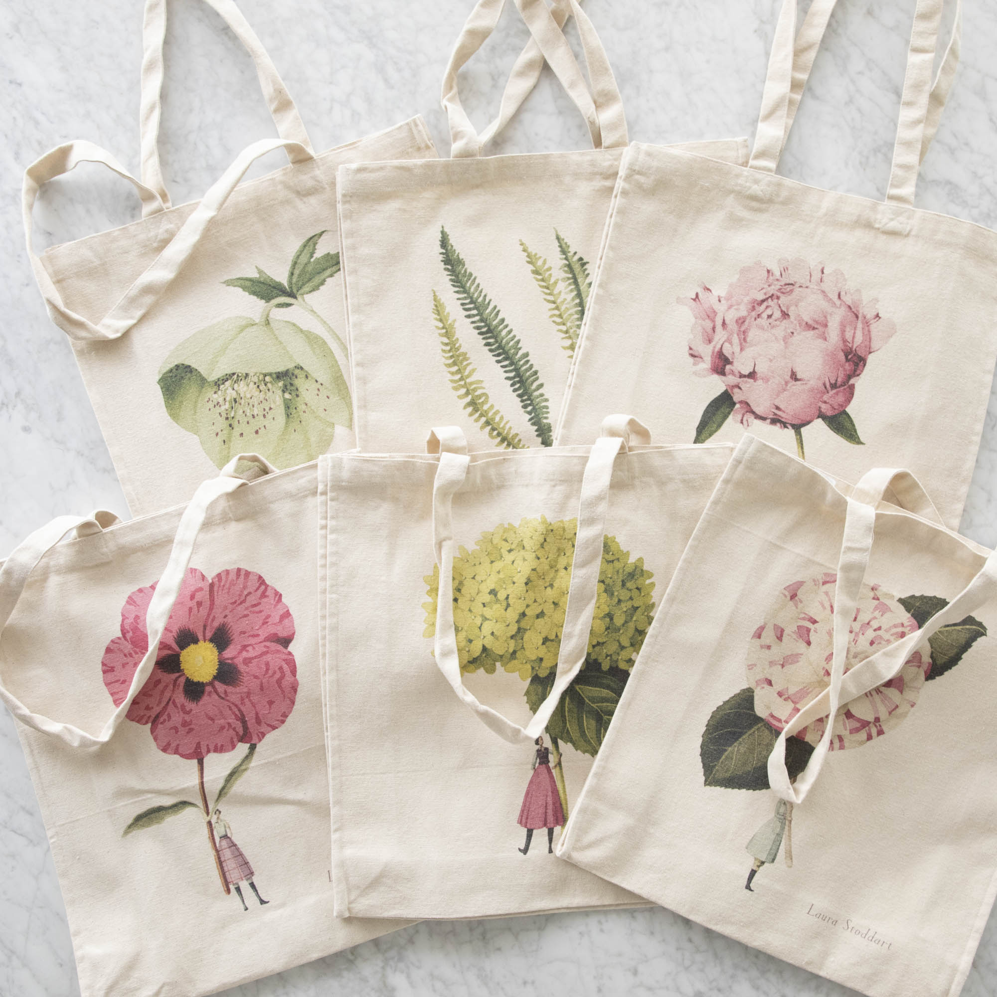 Six of the Heavyweight Canvas Tote Bags featuring different illustrations of small people holding large plants by the stem by Laura Stoddart, laying flat and overlapping on a white marble table.