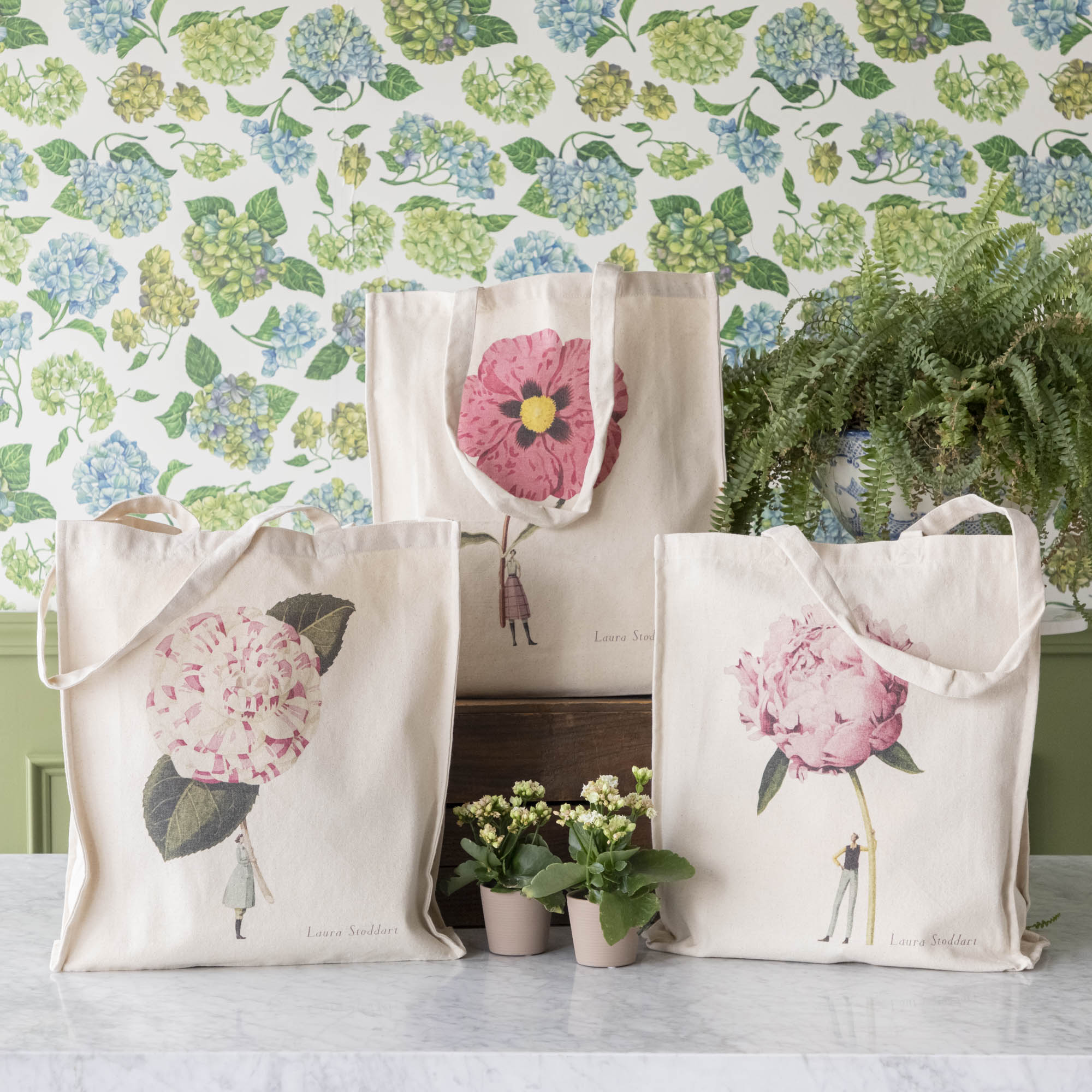 Three of the Heavyweight Canvas Tote Bags featuring pink floral artwork by Laura Stoddart arranged alongside potted plants against a hydrangea wallpaper background.