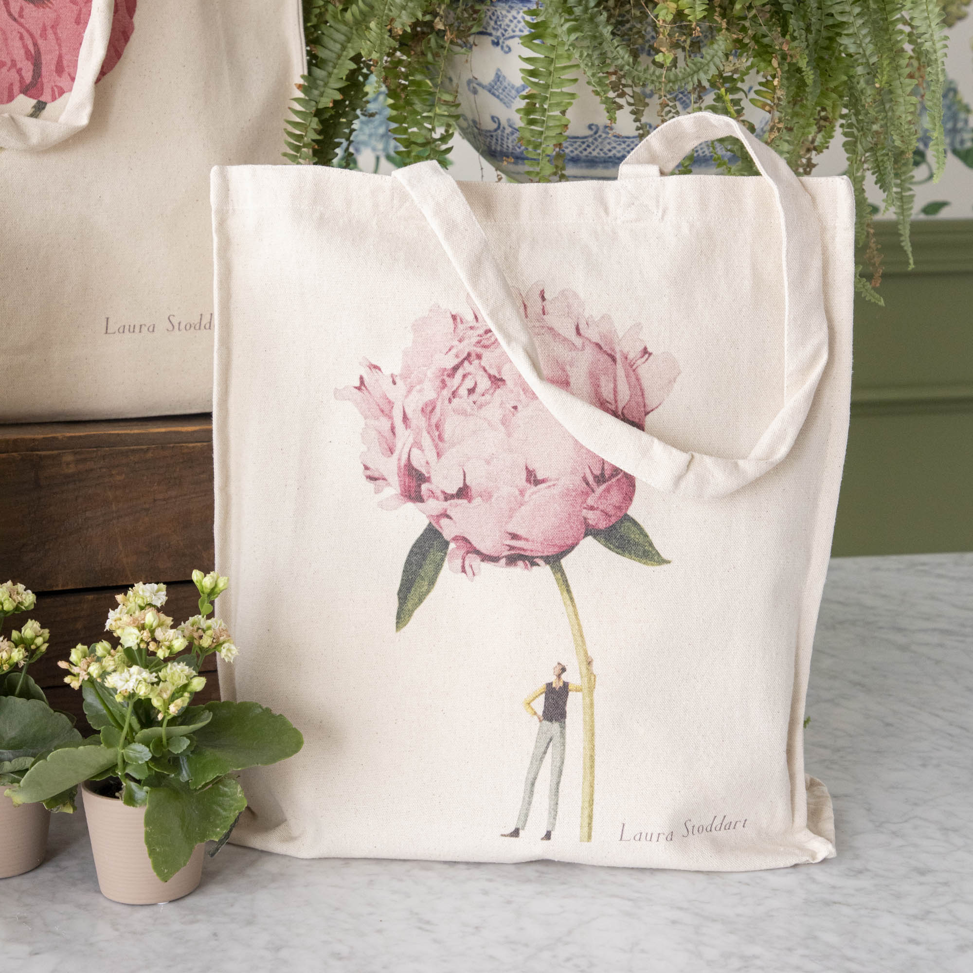 Close-up of the Pink Peony Heavyweight Canvas Tote Bag standing upright in a display, showing the beautiful illustration of a man holding a gigantic pink flower.