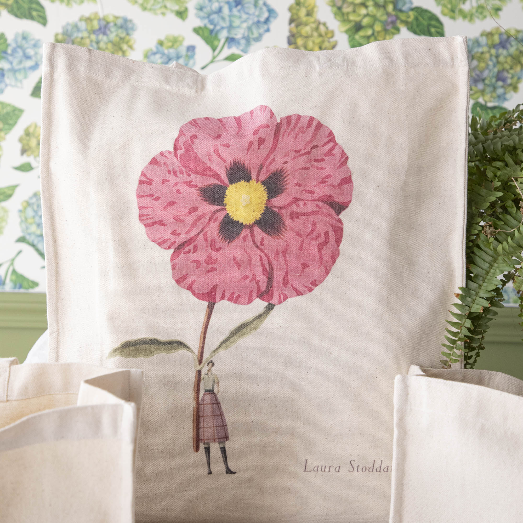 Close-up of the Cistus Heavyweight Canvas Tote Bag standing upright in a display, showing the beautiful illustration of a woman holding a gigantic pink flower.