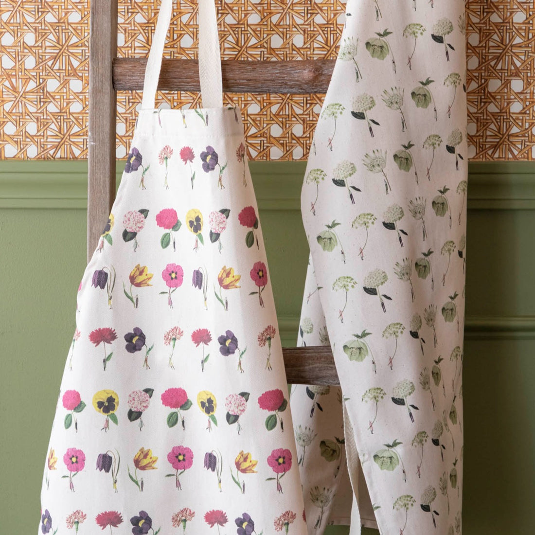 Two Laura Stoddart Aprons by Hester &amp; Cook hanging on a wooden ladder against a wall with patterned wallpaper above wainscoting.