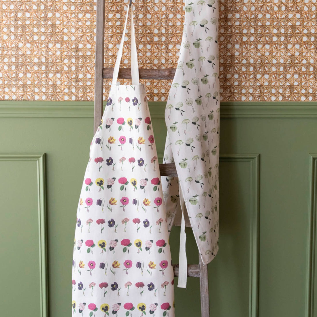 Two Laura Stoddart Aprons by Hester &amp; Cook hanging on a wooden ladder against a wall with patterned wallpaper above wainscoting.