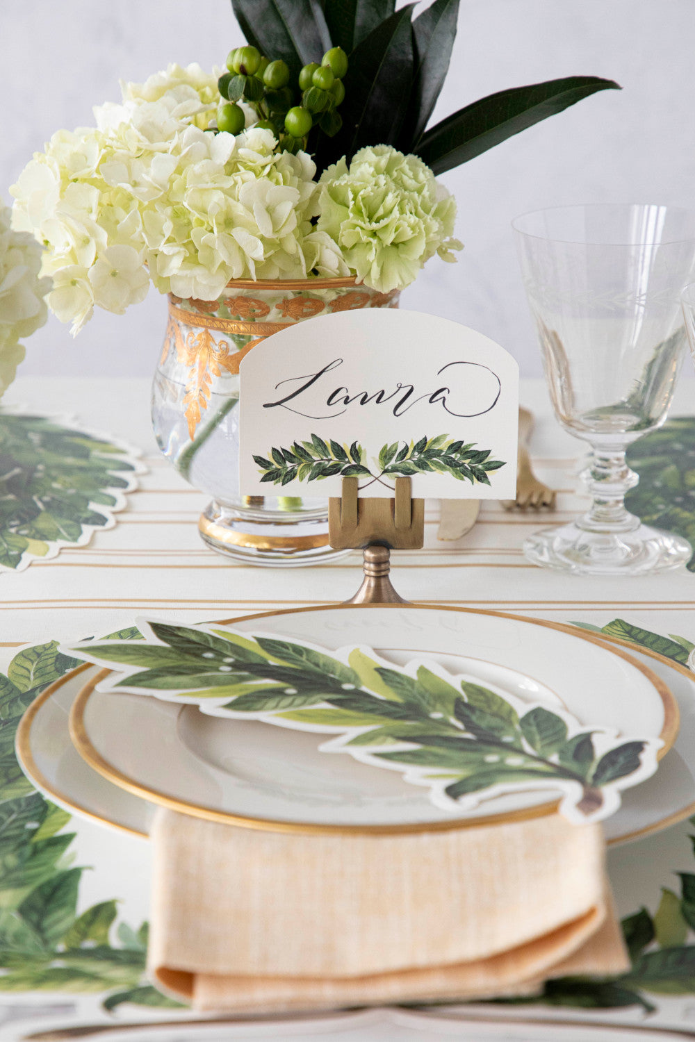 An elegant place setting featuring a Laurel Place Card labeled &quot;Laura&quot; in a brass place card holder behind the plate.