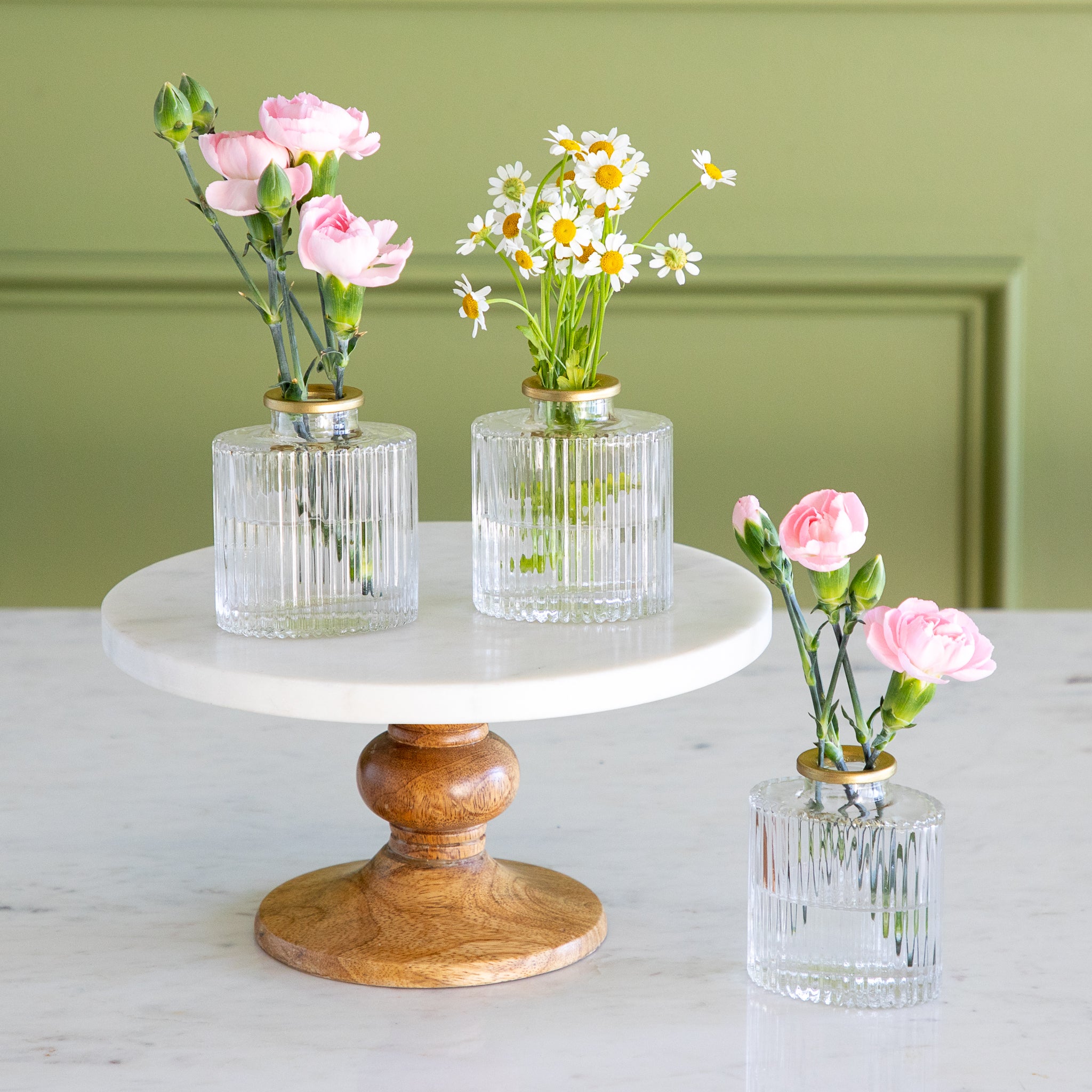Three glass vases with flowers on a marble stand against a green wall.