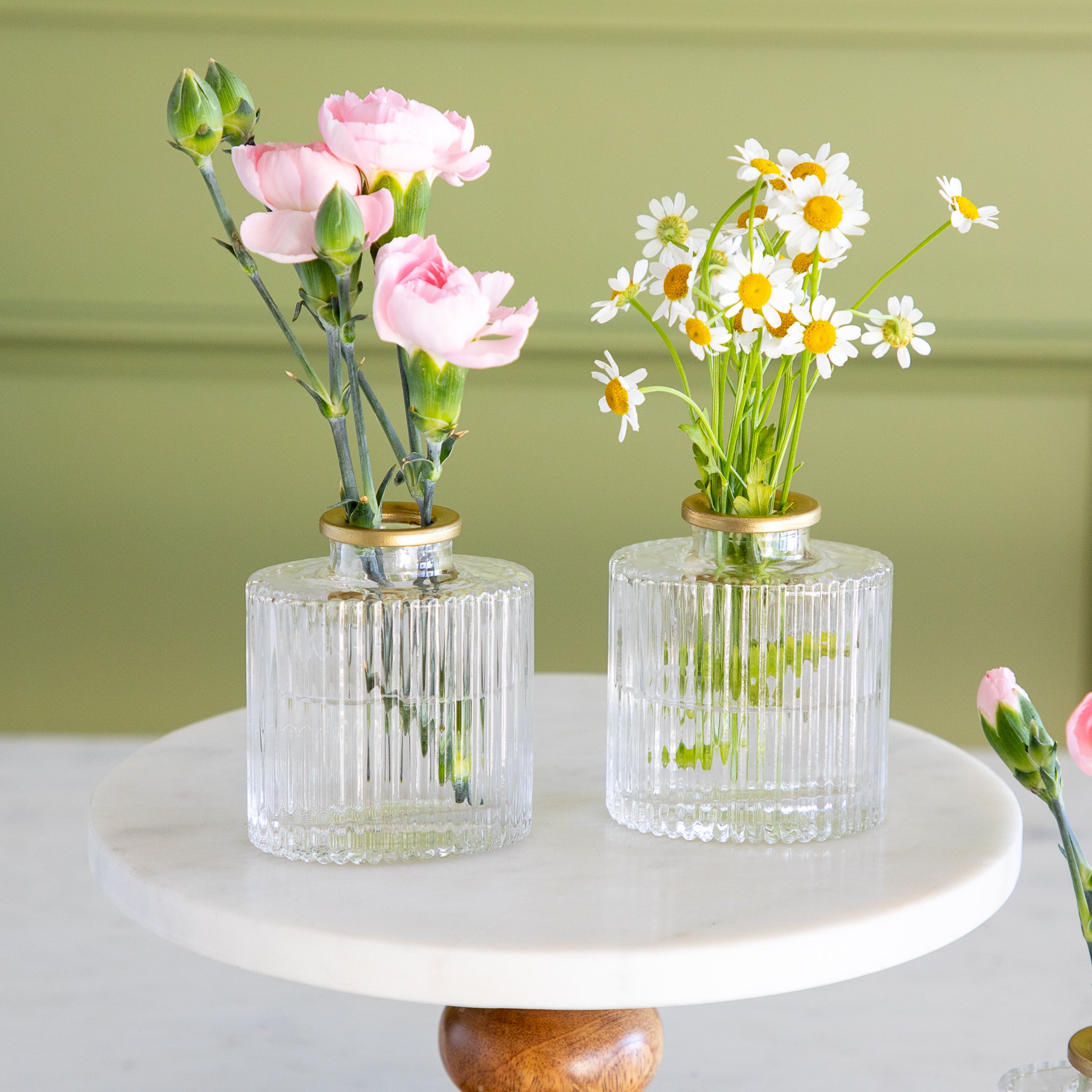 Two glass vases with floral arrangements on a white surface against a green background.