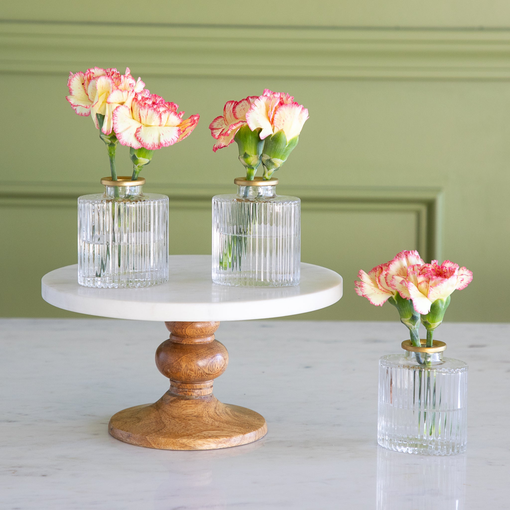 Three small vases with flowers on a marble and wood stand against a green wall.