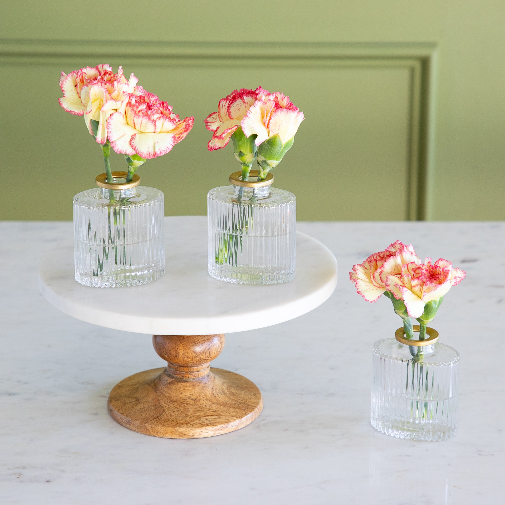 Three small glass vases with flowers on a marble surface and wooden stand against a green wall.