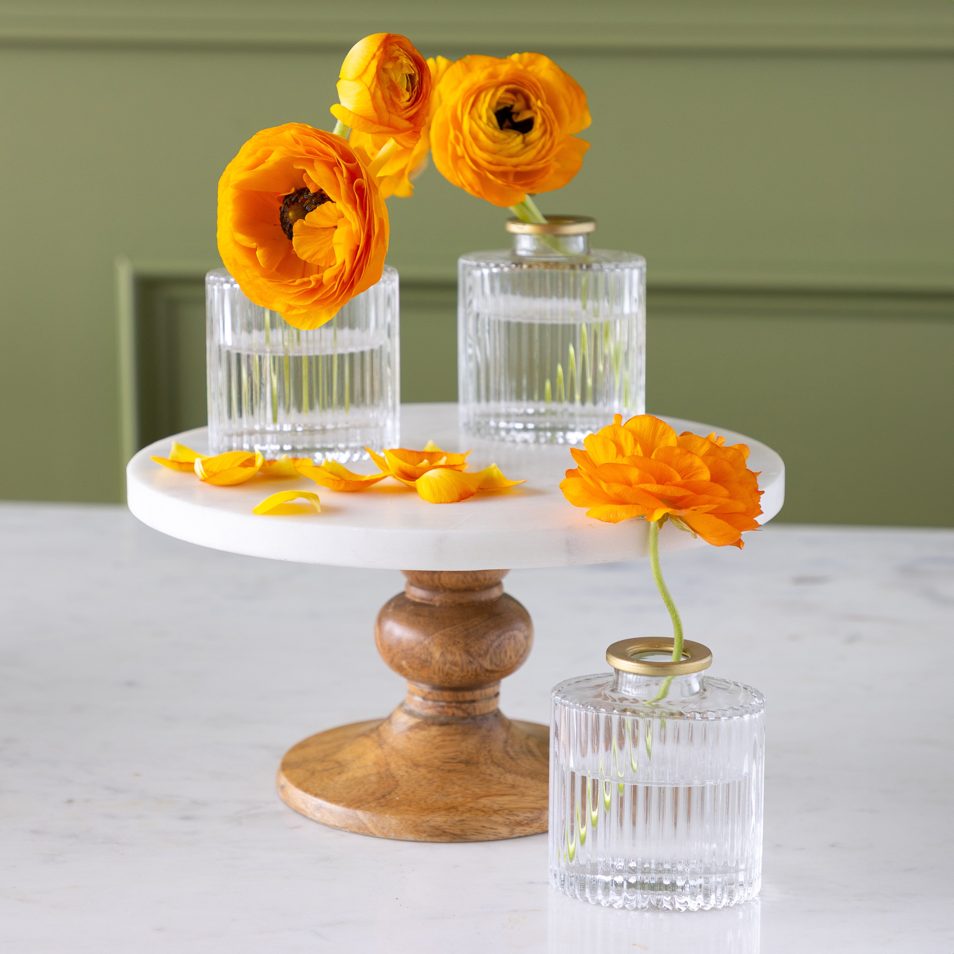 Three Petite Gold Rim Oval Bud Vases with orange ranunculus flowers and scattered petals are arranged on a round marble and wood cake stand, set against a green wall.