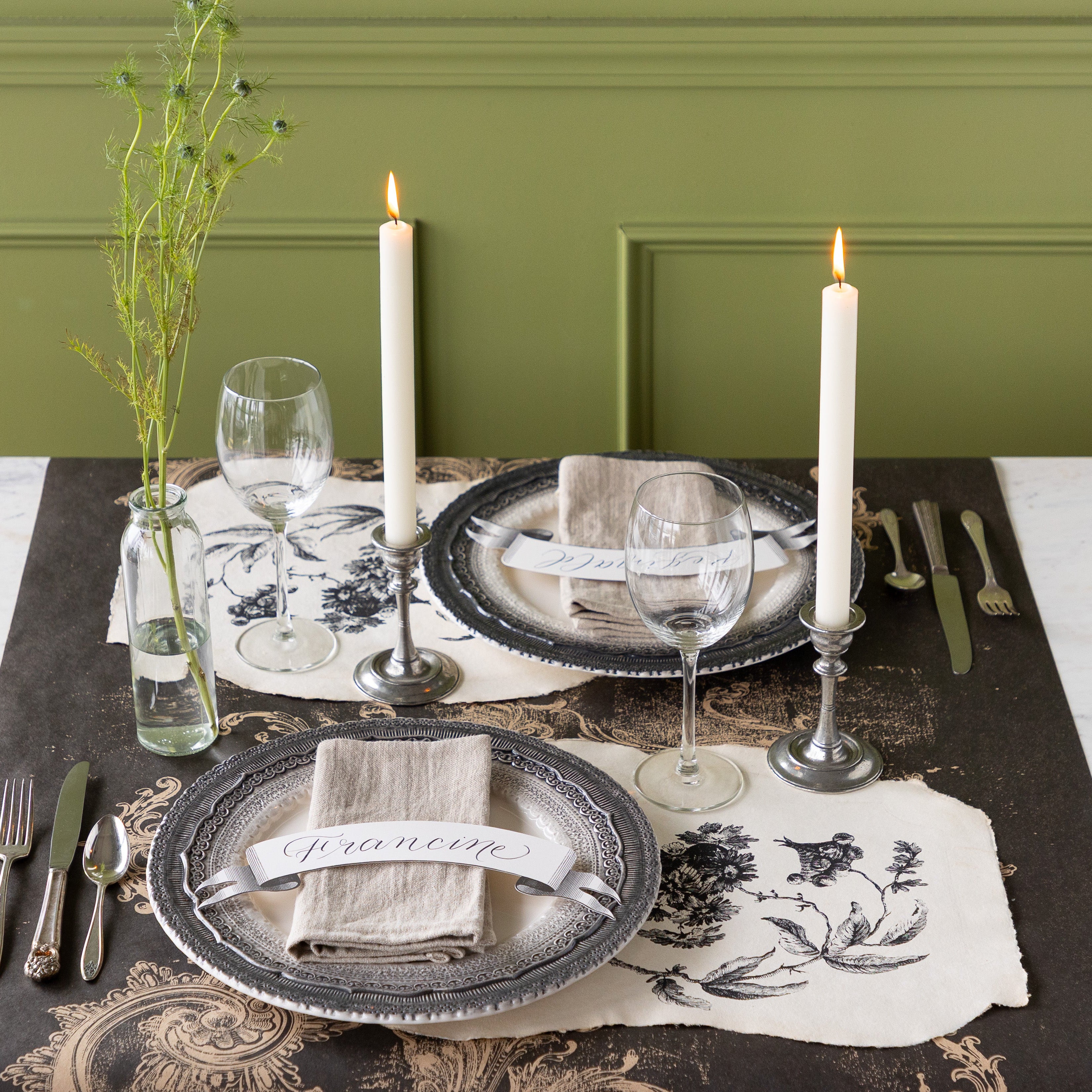 Table setting with decorative plates, Pillement Paper Placemats, glasses, and candles on the Black Archival Runner against a green wall.