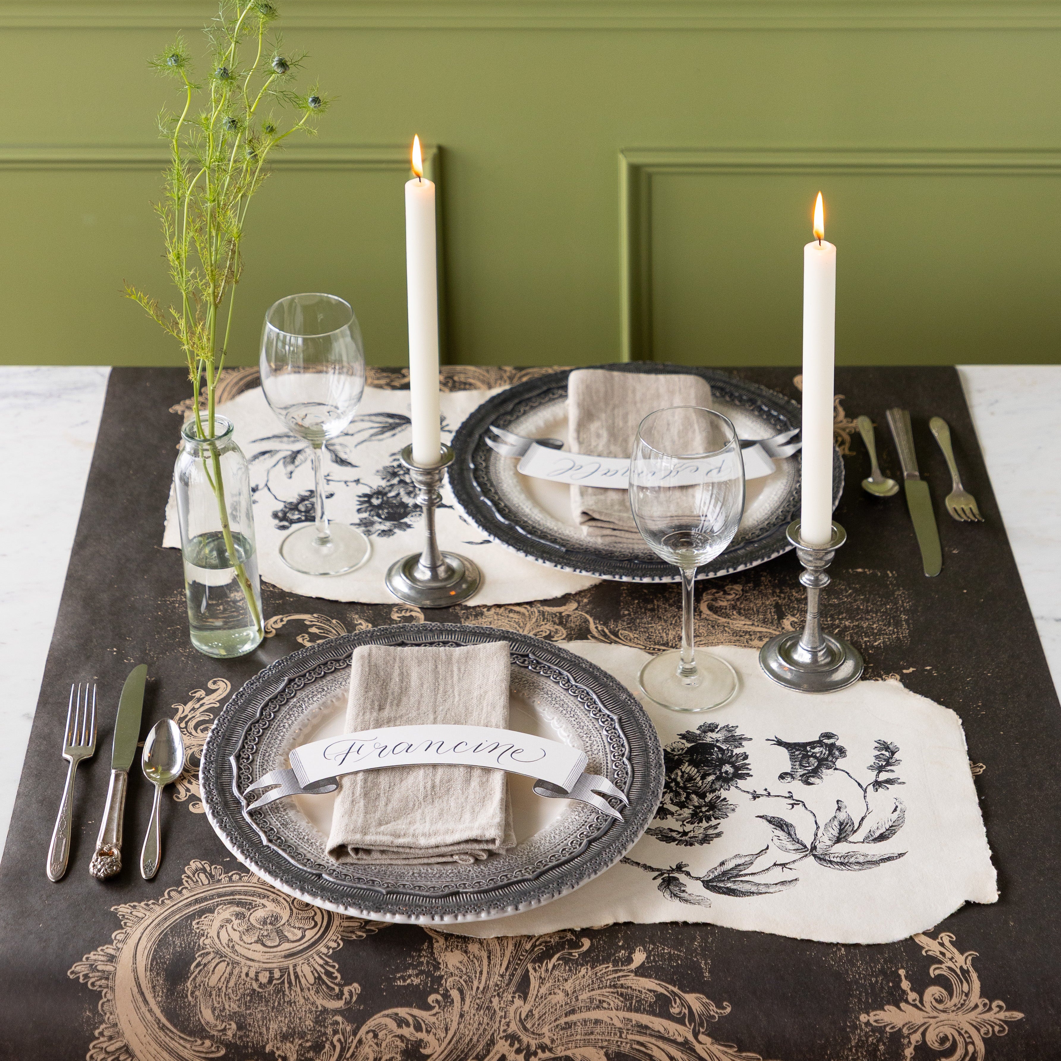 Table setting with decorative plates, Pillement Paper Placemats, glasses, and candles on the Black Archival Runner against a green wall.