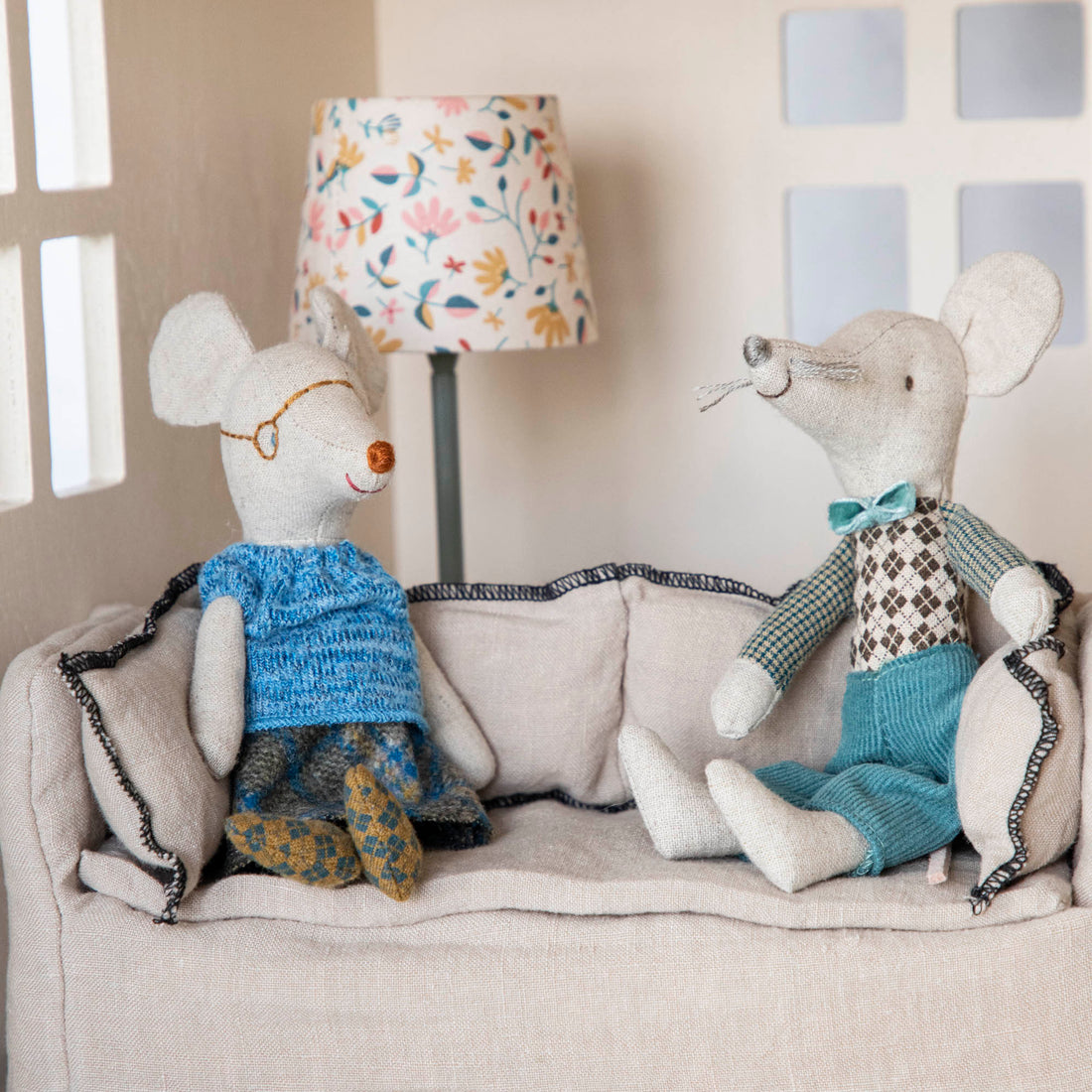 Two plush Maileg mouse toys, resembling Grandma and Grandpa Mice, seated on a miniature couch beside a small lamp.
