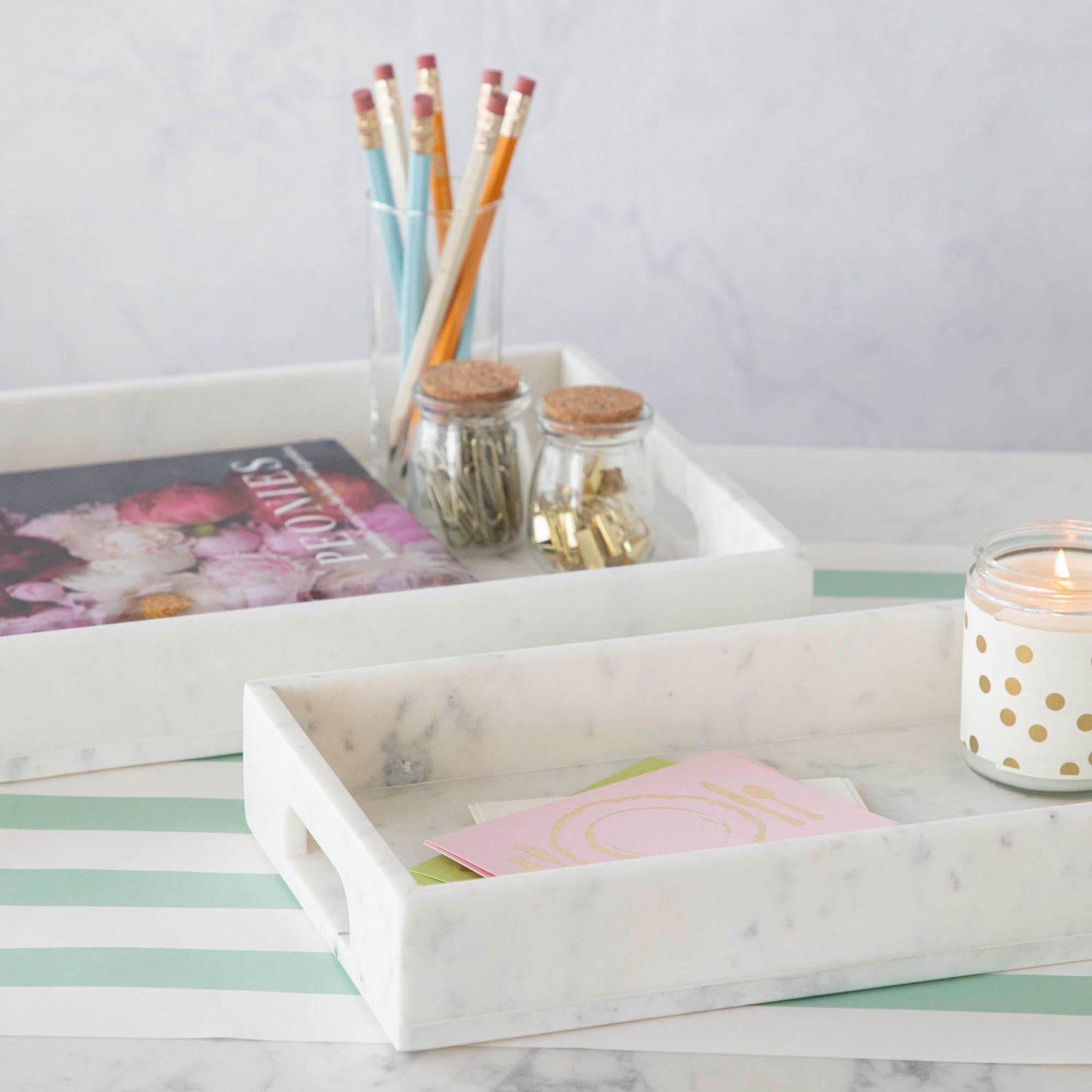 Two Godinger White Marble Rectangular Trays on a striped surface, one holding art supplies and the other candles and a small jar, showcasing the elegance of marble.