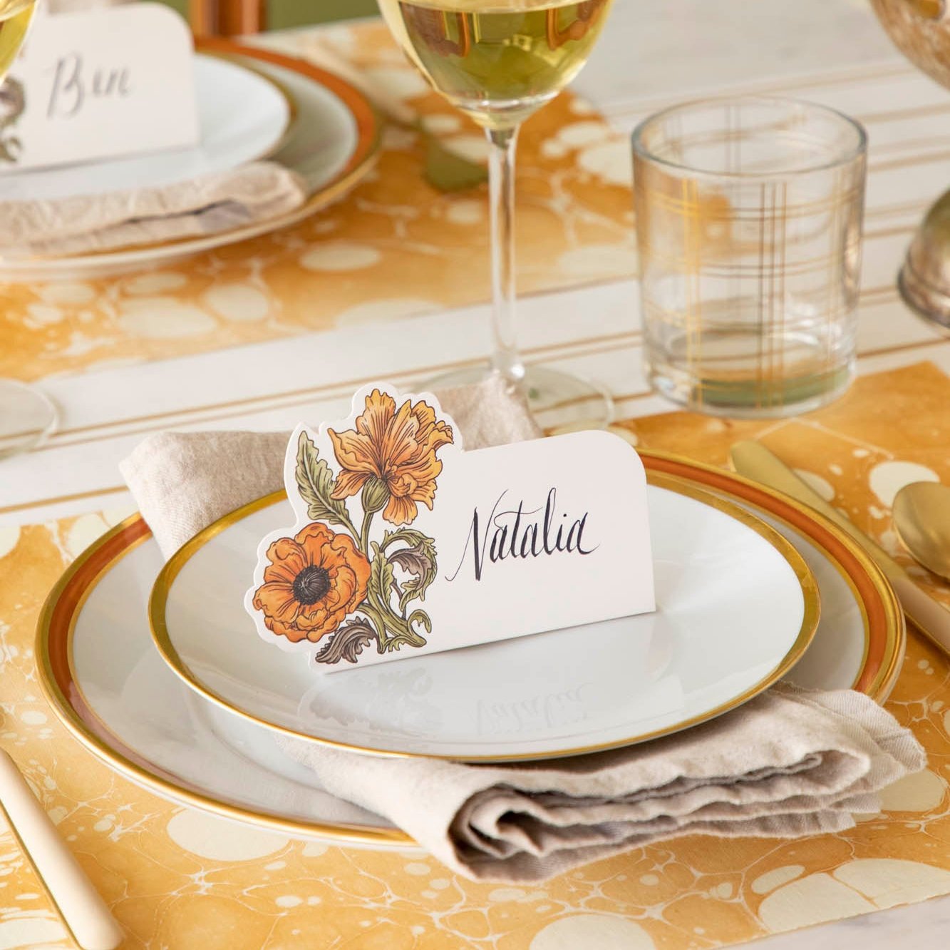 The Autumn Arrangement Place Card with the name "Natalia" written on it, on an elegant gold themed table setting.