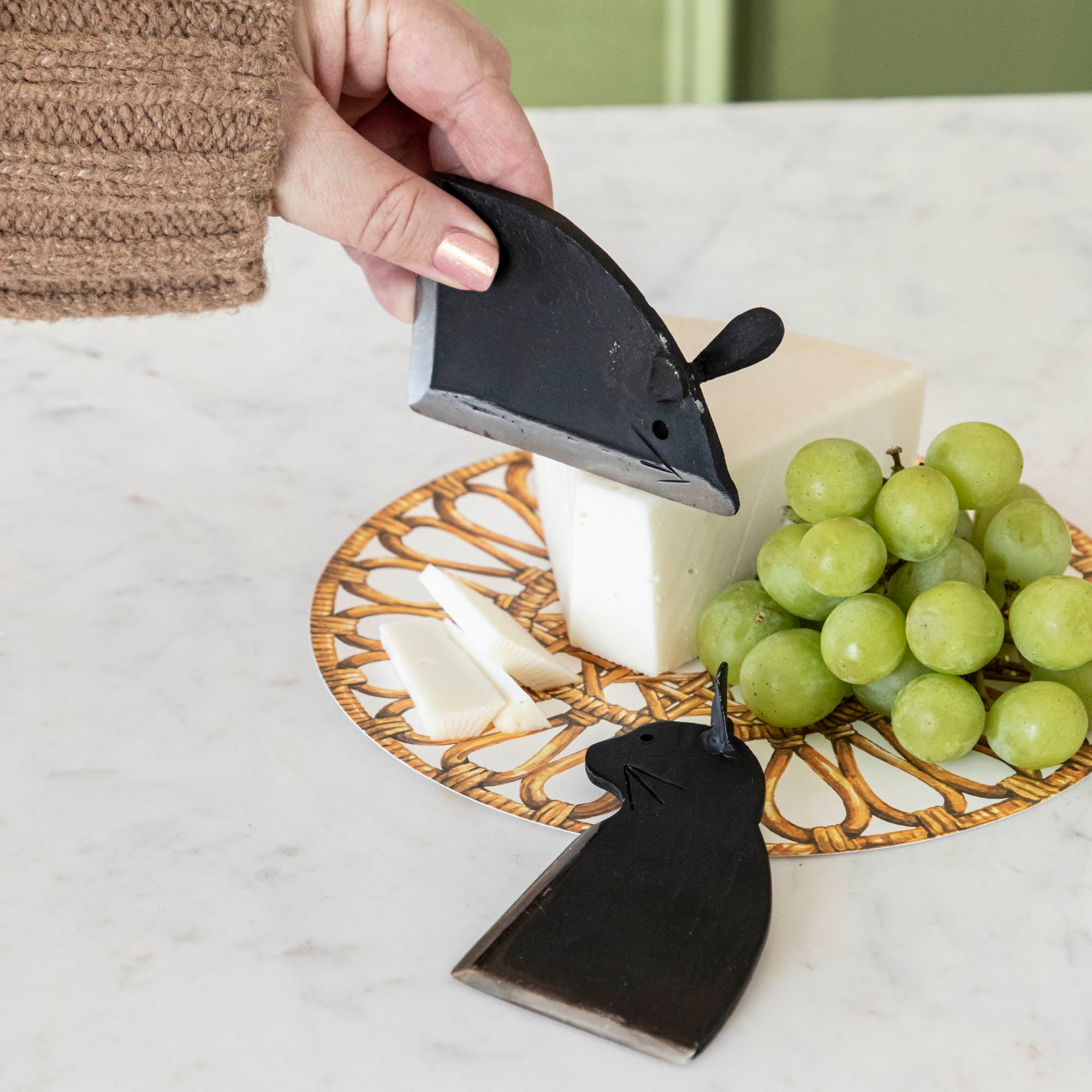 Cheese and grapes on a plate with a cheese knife shaped like a mouse and a cat on a marble surface.