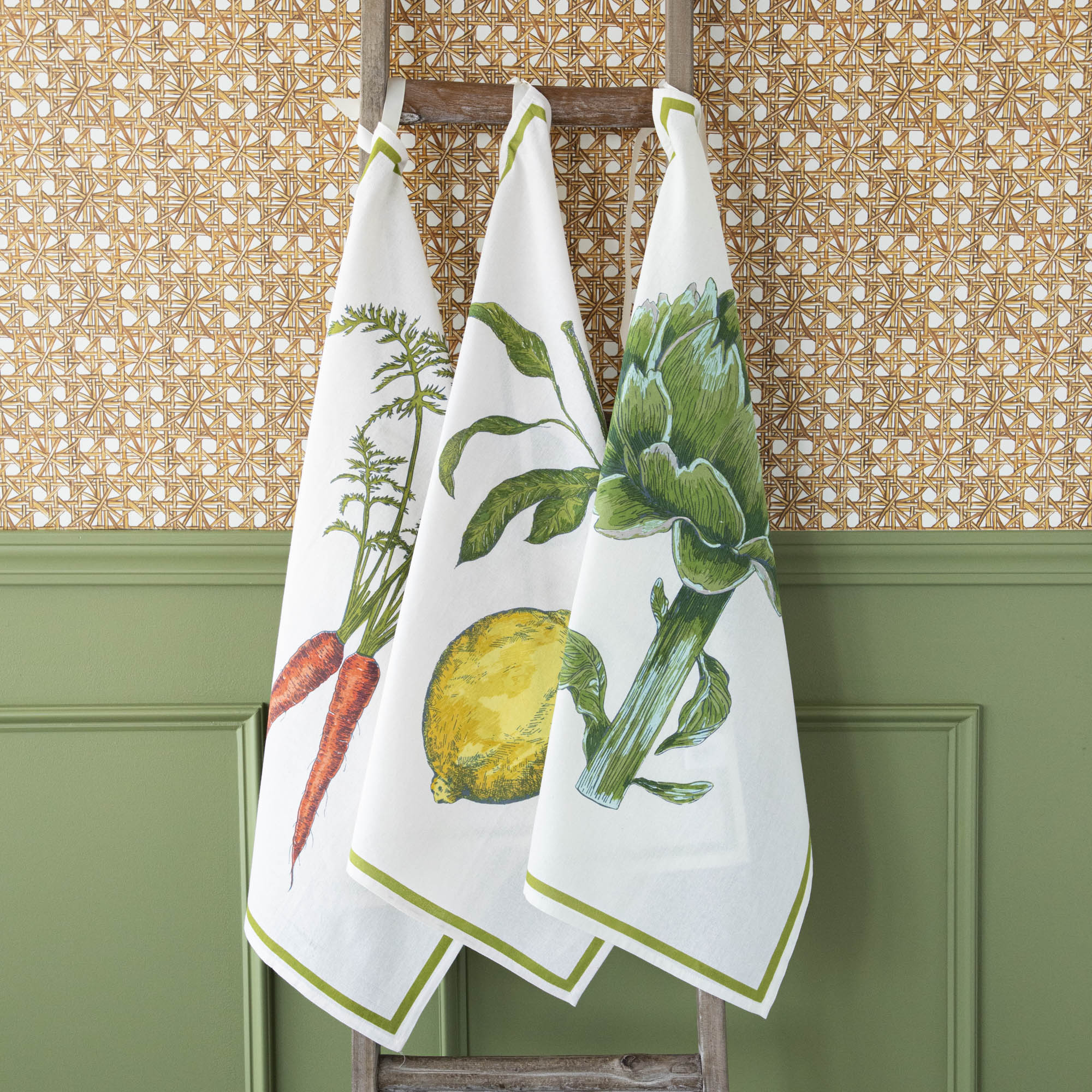 A Lemon Flour Sack Kitchen Towel Set of 2 with a lemon design hanging on a wooden deck railing, with a bowl of lemons in the background. (Mahogany)