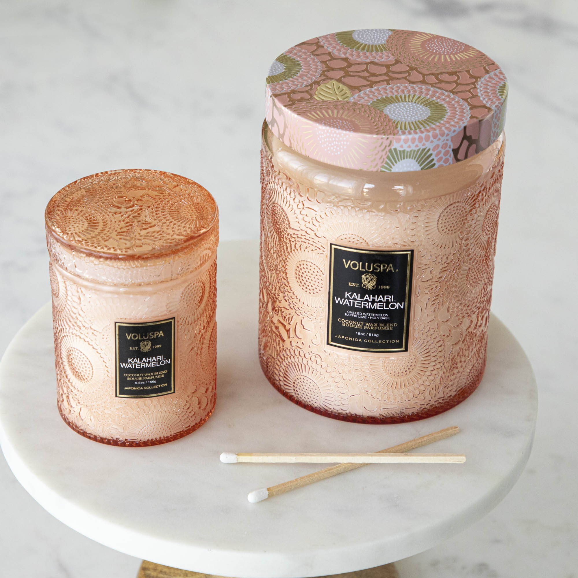 Two Voluspa Kalahari Watermelon candles on a marble surface with matches nearby, featuring a crisp cucumber aroma.