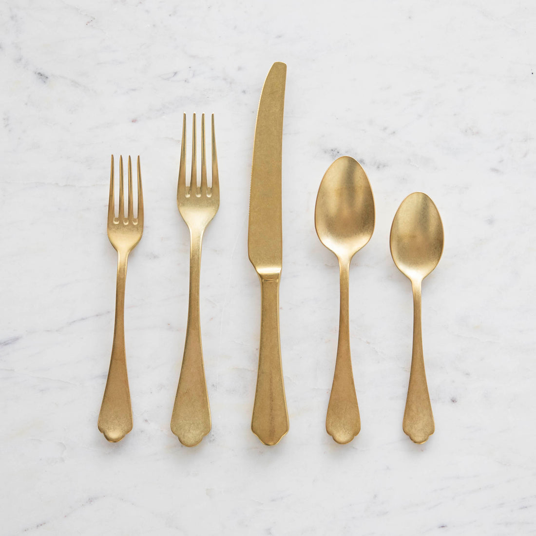 A set of Mepra Dolce Vita Pewter Oro 5-Piece Place Setting flatware, including two forks, a knife, and two spoons, neatly arranged on a marble surface.