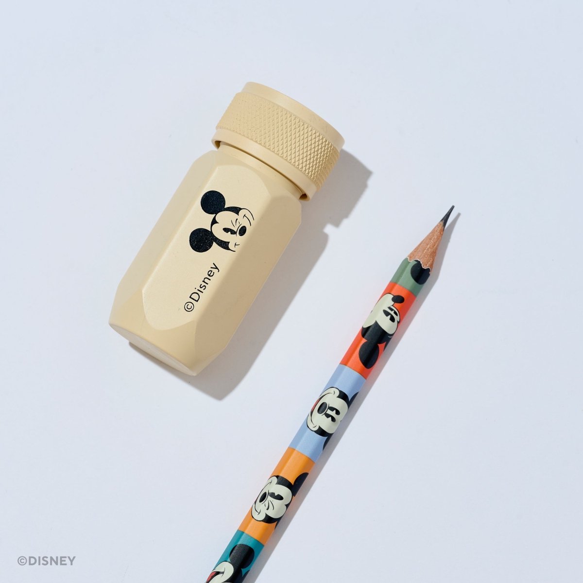 A Disney pencil and matching pencil sharpener set on a light gray background. The sharpener is a light beige plastic hexagonal tool with a black and white Mickey Mouse graphic and the Disney name.