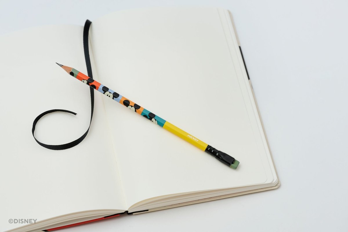 A yellow pencil with a colorful Mickey Mouse pattern rests on an open blank notebook with a black ribbon bookmark, shot on a clean white background.
