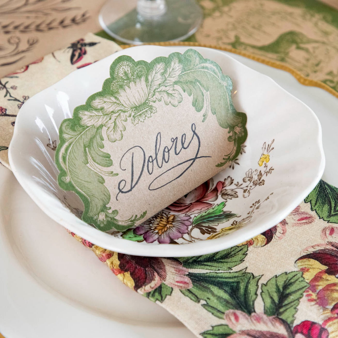 An elegant place setting featuring a Moss Fable Toile Place Card labeled &quot;Dolored&quot; standing in the bowl.