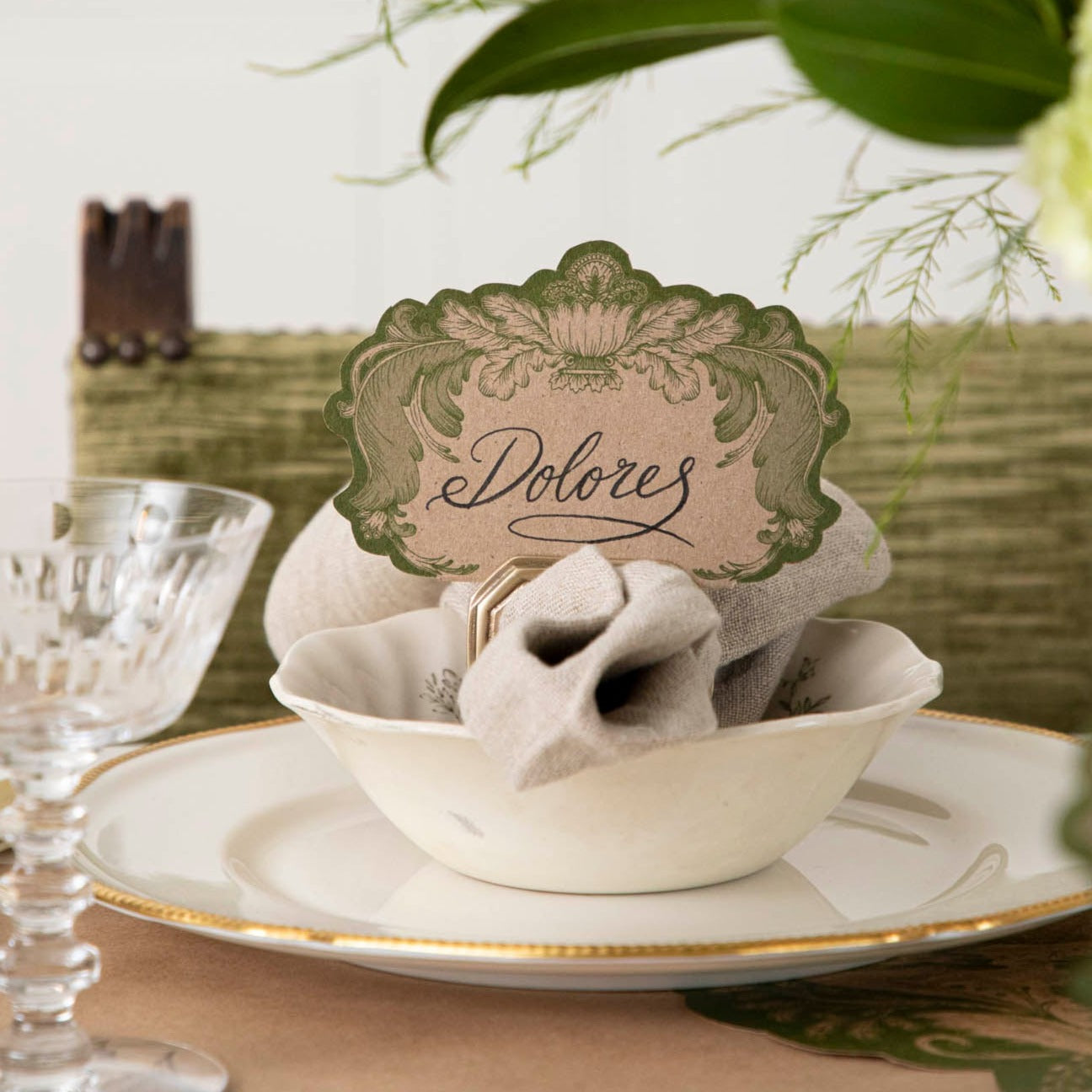 A Moss Fable Toile Place Card labeled &quot;Dolored&quot; standing in a napkin ring place card holder, as part of an elegant table setting.